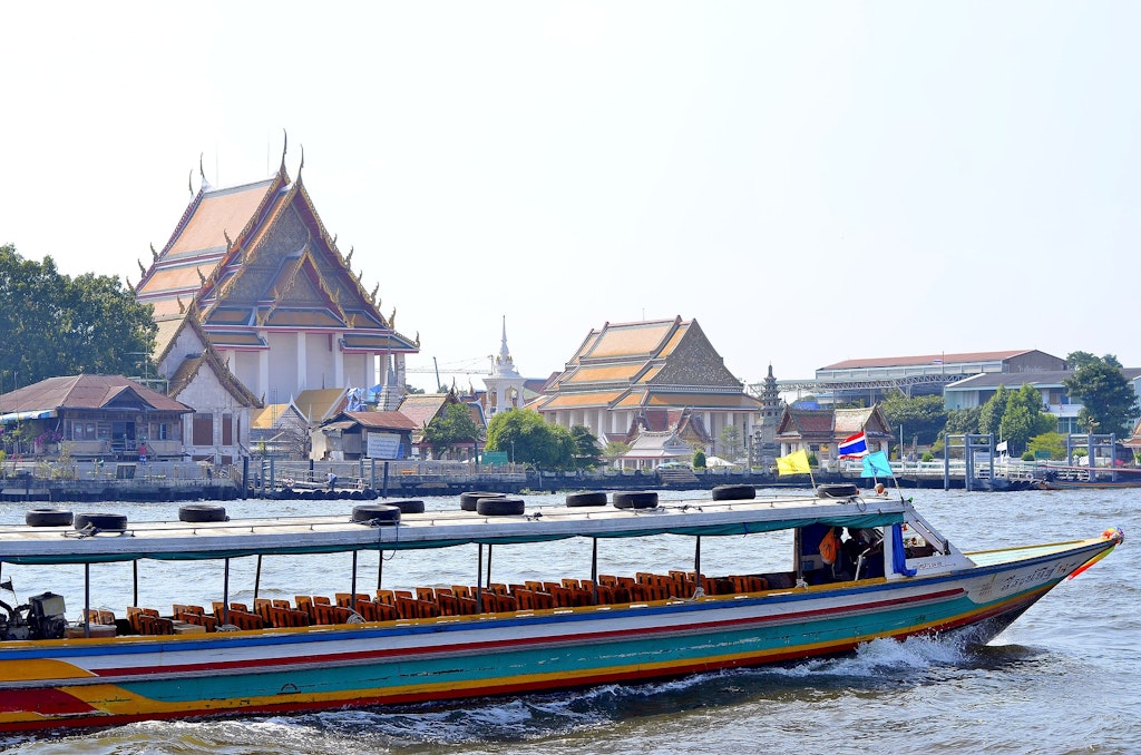 An empty boat floats on the Chao Phraya River in Bangkok, its wooden frame in diverse colors. In the back you can see traditional Thai buildings.