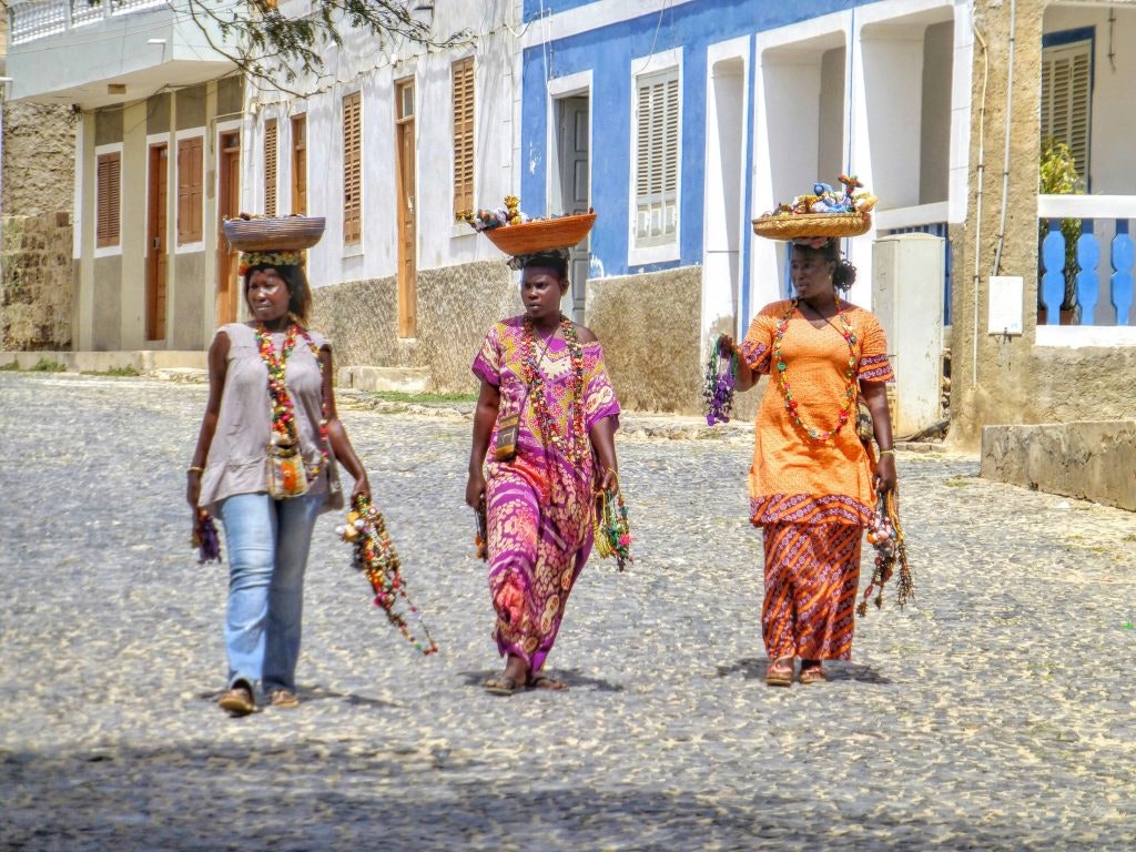 Three women in traditional clothing walking on a cobblestone street with baskets on their heads in Cape Verde