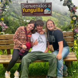 best service trips for teens two girls and one guy sitting on a bench and above them is a sign saying "bienvenidos a Yunguilla"
