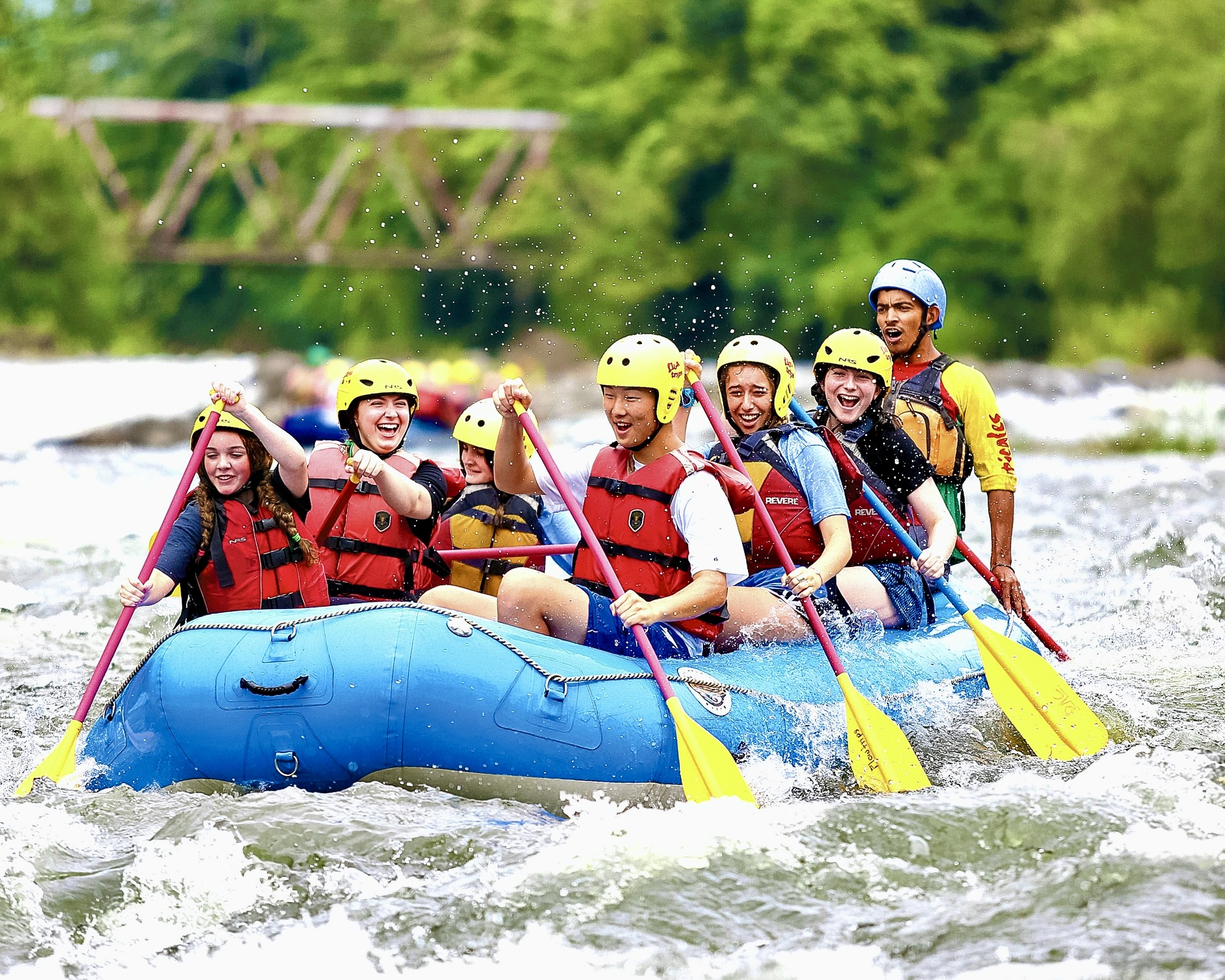 High school students in helmets paddle a whitewater raft through rapids with a guide on a summer adventure camp