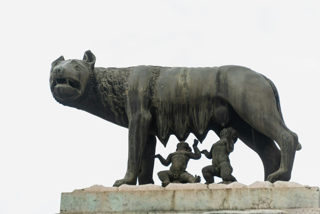 The Capitoline Wolf is a bronze statue of a she-wolf standing on all fours with a fierce expression. She has detailed fur and looks protective. Below her are the two legendary twins, Romulus and Remus.