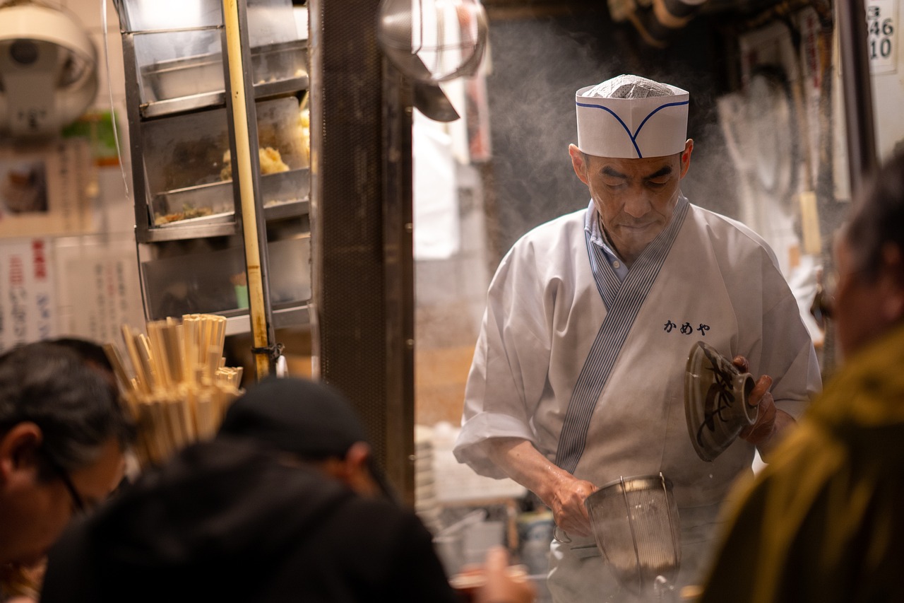 A chef in traditional white attire and cap prepares food at a steamy street food stall