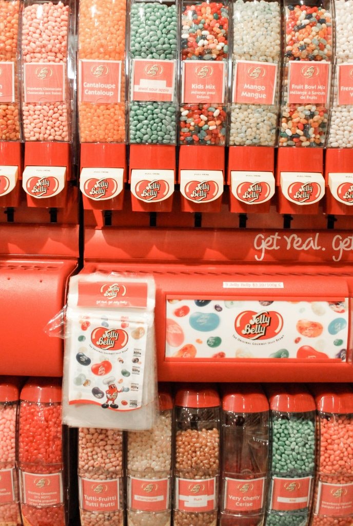 The image shows a colorful Jelly Belly display featuring multiple dispensers with flavors like "Cantaloupe," "Kids Mix," and "Mango".