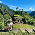 Photo of a person sitting on a rock path on the grass, looking at Ciudad Perdida with blue sky