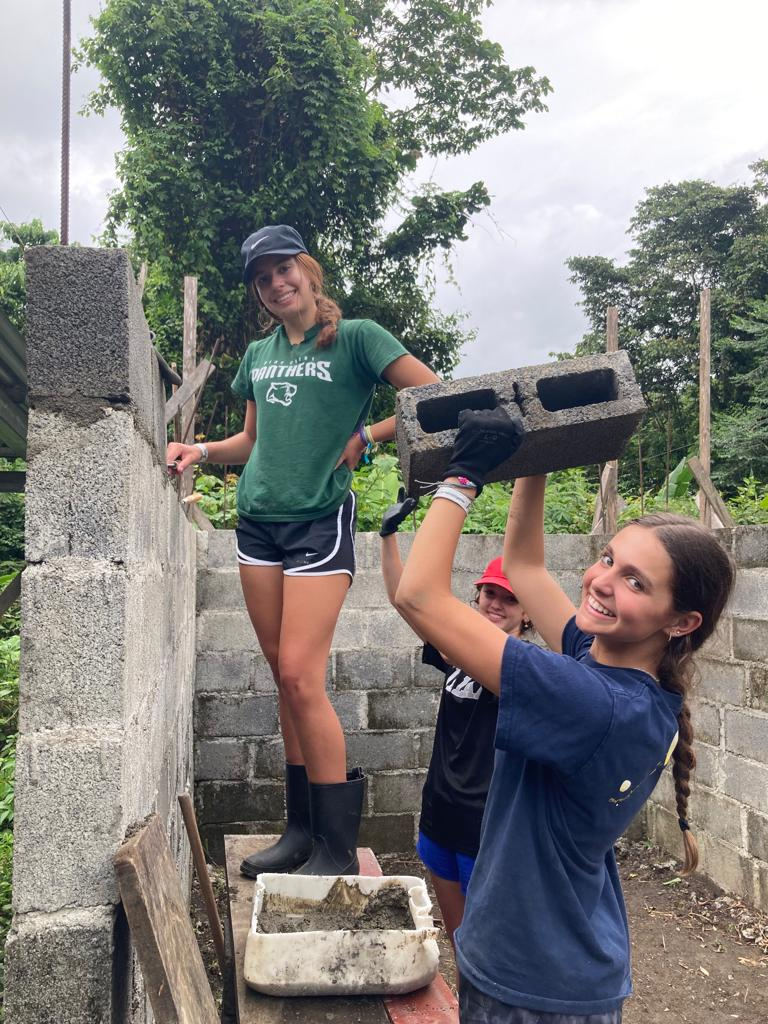2 girl students seen working on a brick wall, one standing on top of something smiling agains the wall while the other in front holding up a big brick while smiling.