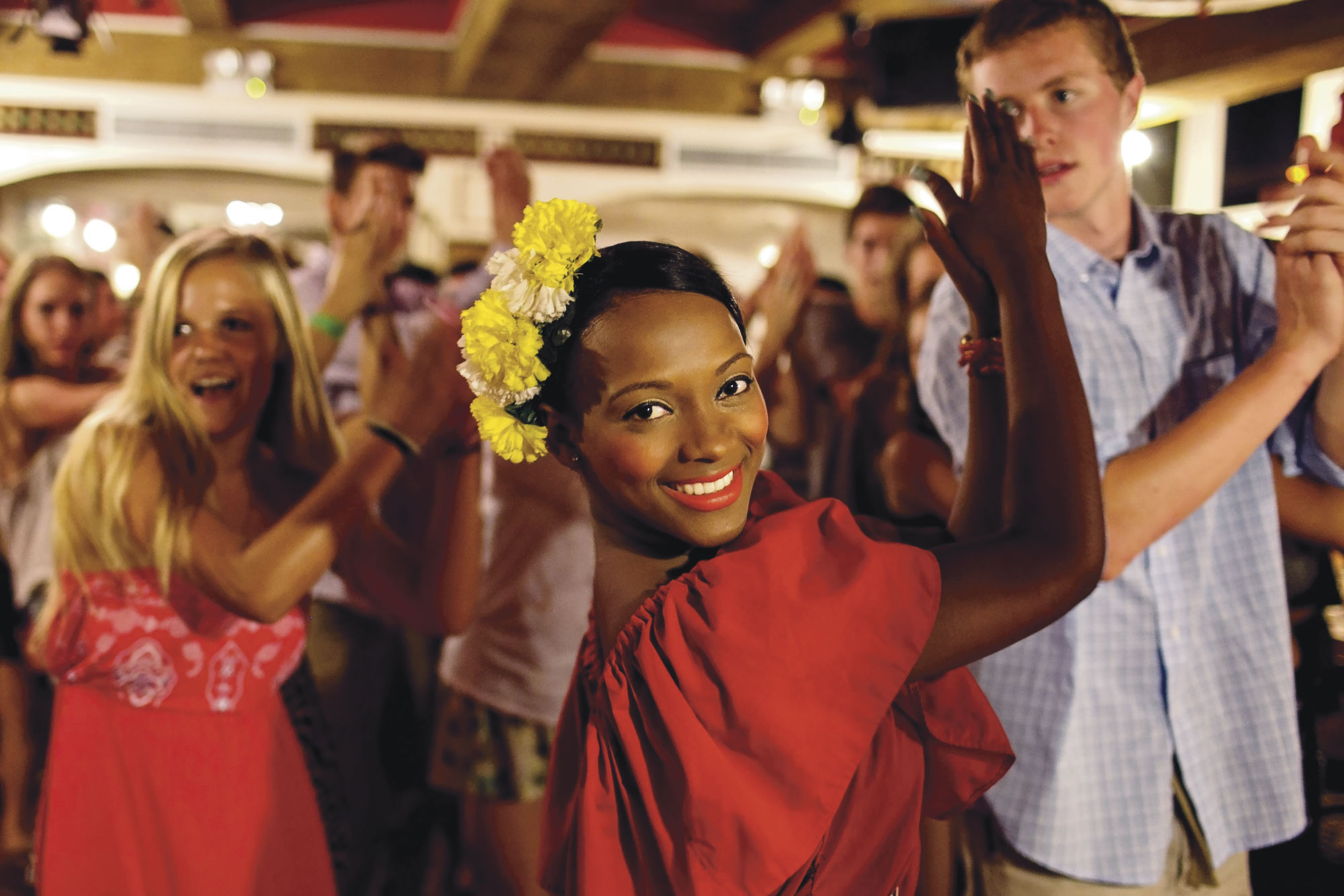 Students dance indoors, one wearing a flower crown and red dress