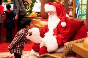 Santa sits on a seat and talks to a young boy who is leaning towards him.