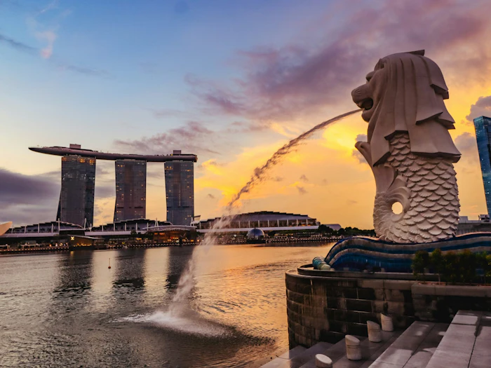The Merlion statue spouts an arc of water into Marina Bay at sunset, with the three towers of Marina Bay Sands on the far bank.