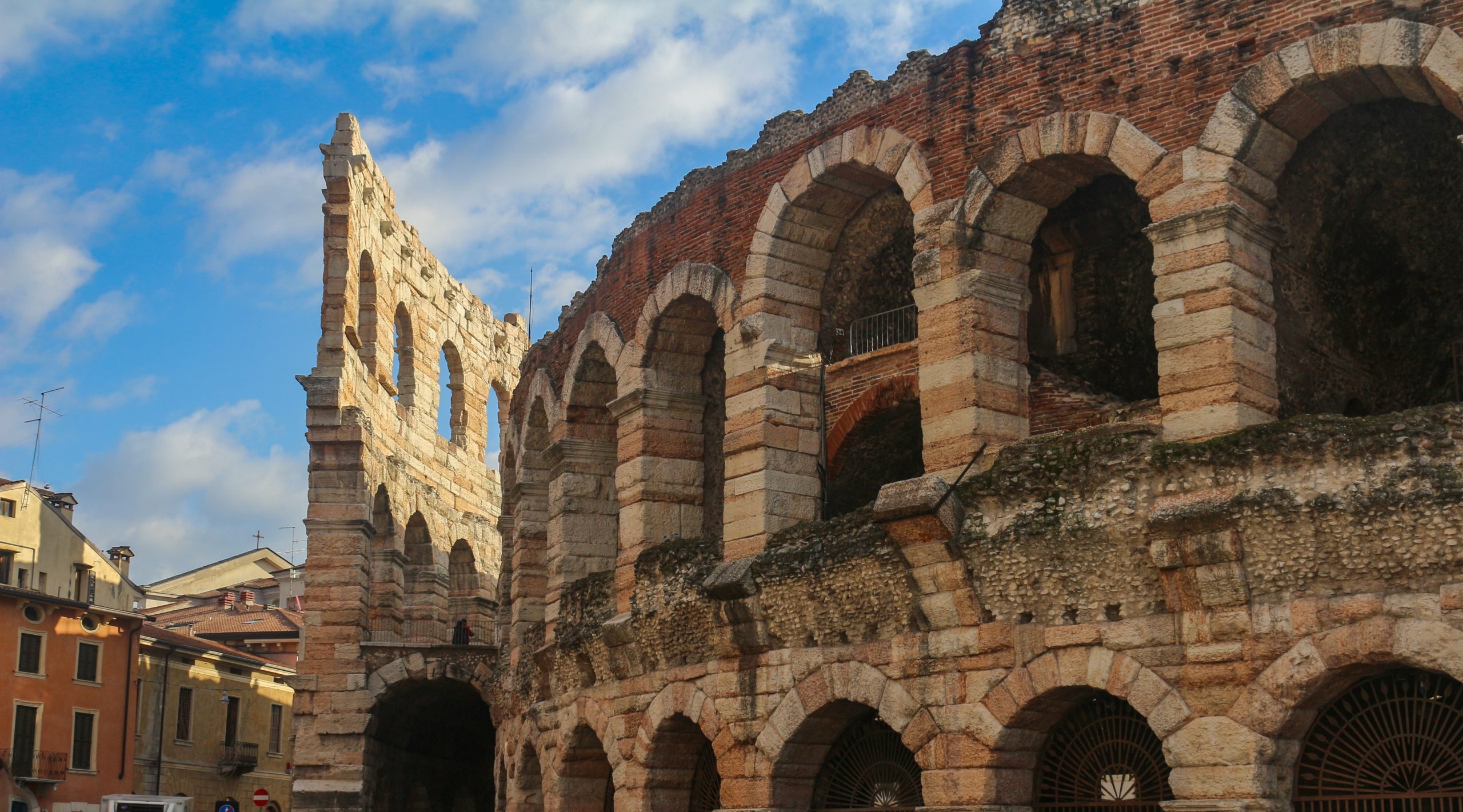 Students walk beneath the weathered stone arches of the Verona Arena under a blue sky