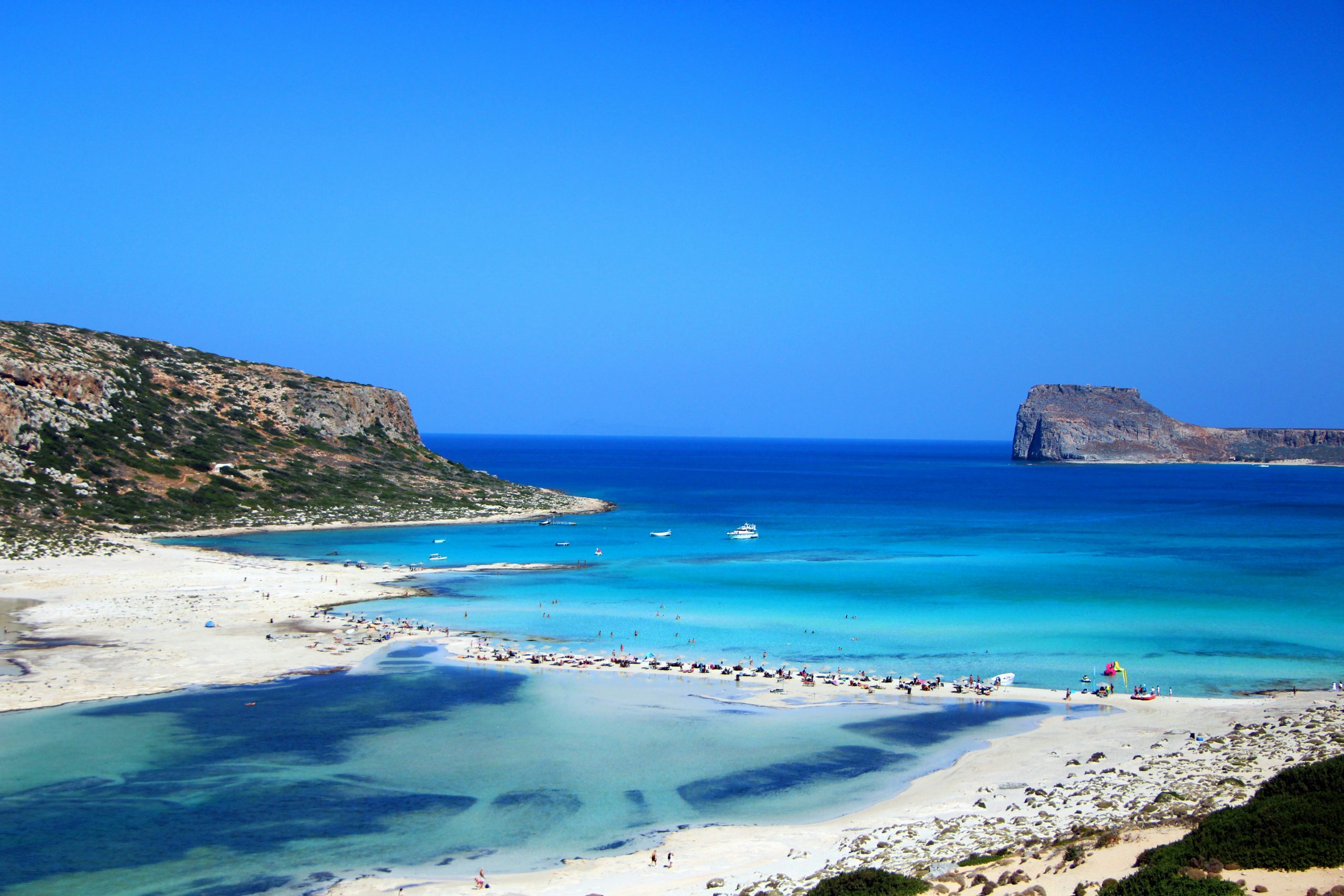A turquoise lagoon behind a pale sandbar, with a rocky headland on one side and a flat-topped island offshore.