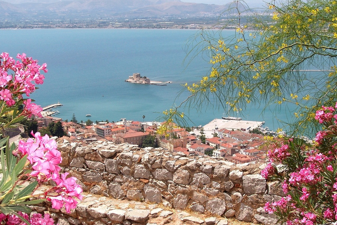 A school trip stops at a stone fortress wall overlooking a coastal town and harbor, framed by pink and yellow blossoms