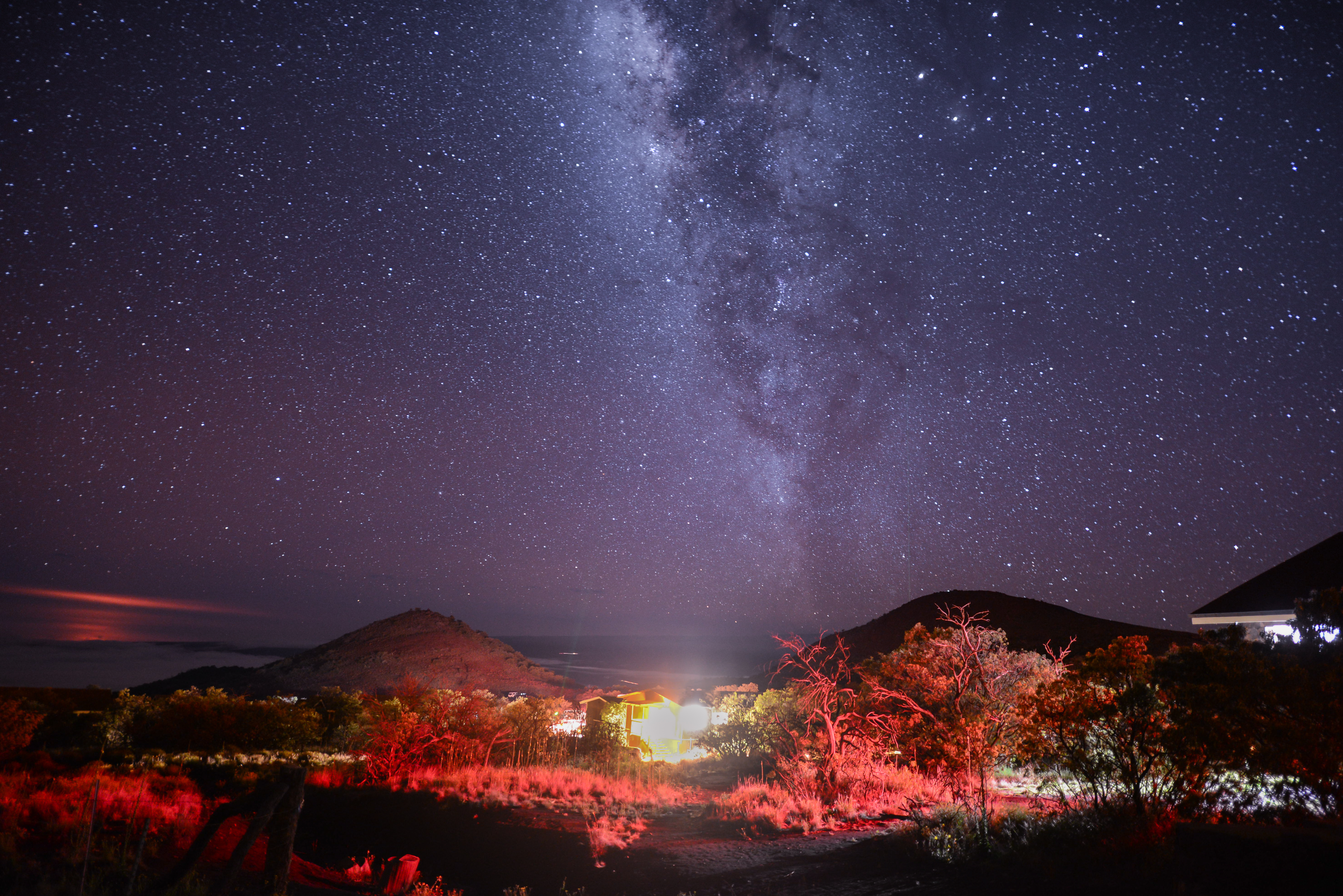 The Milky Way stretches over dark hills with a glowing campsite below