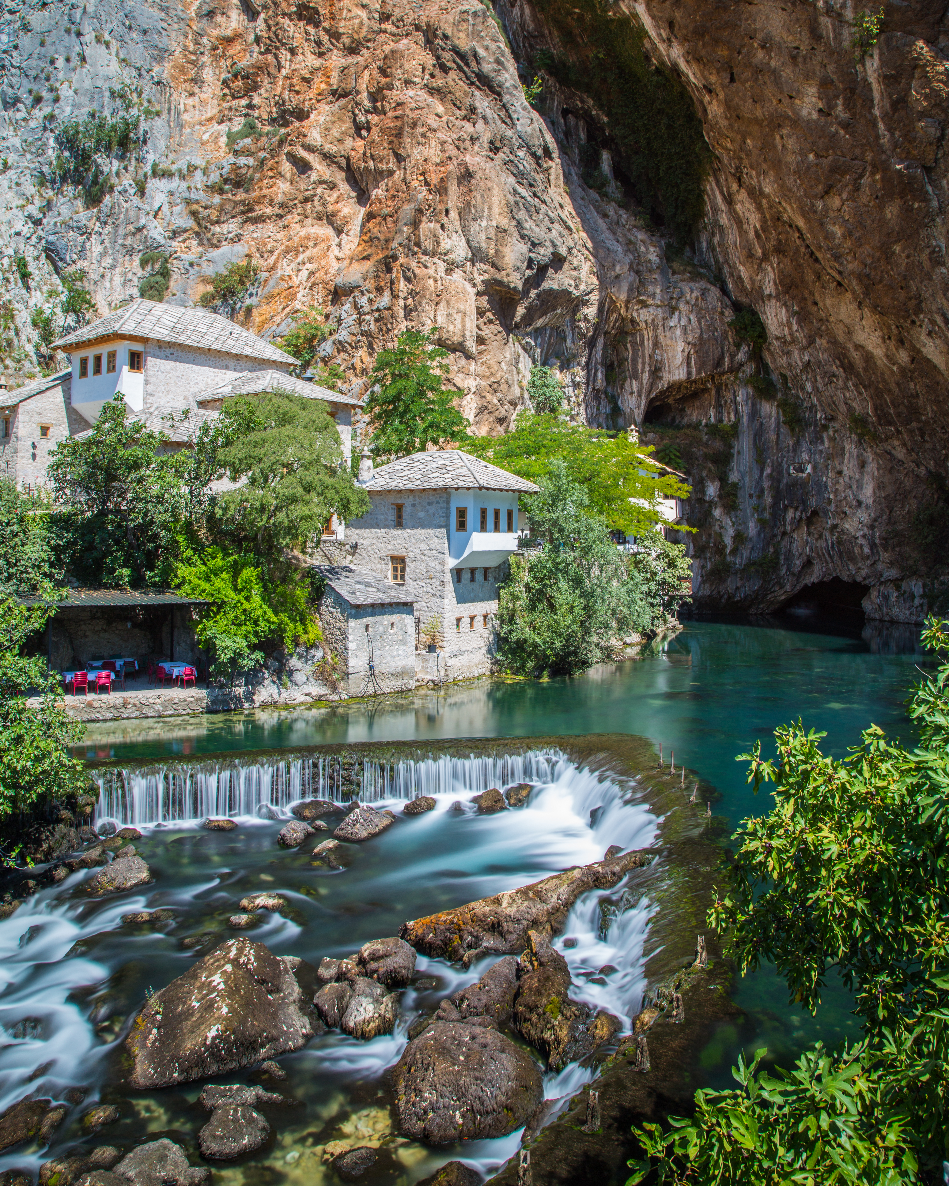 A stone house sits beside a turquoise river emerging from a cliffside cave in Bosnia