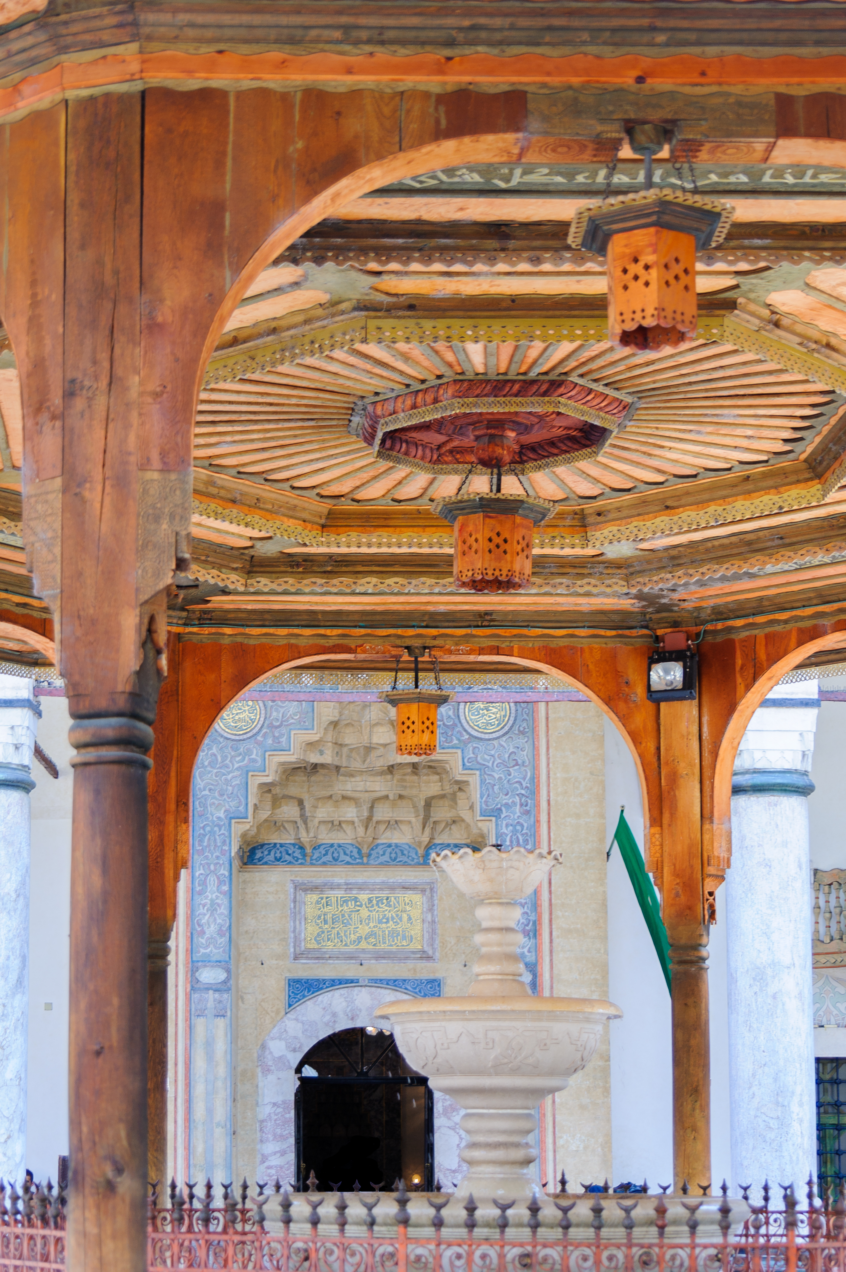 Carved wooden pavilion roof and lanterns over a stone fountain at a mosque courtyard