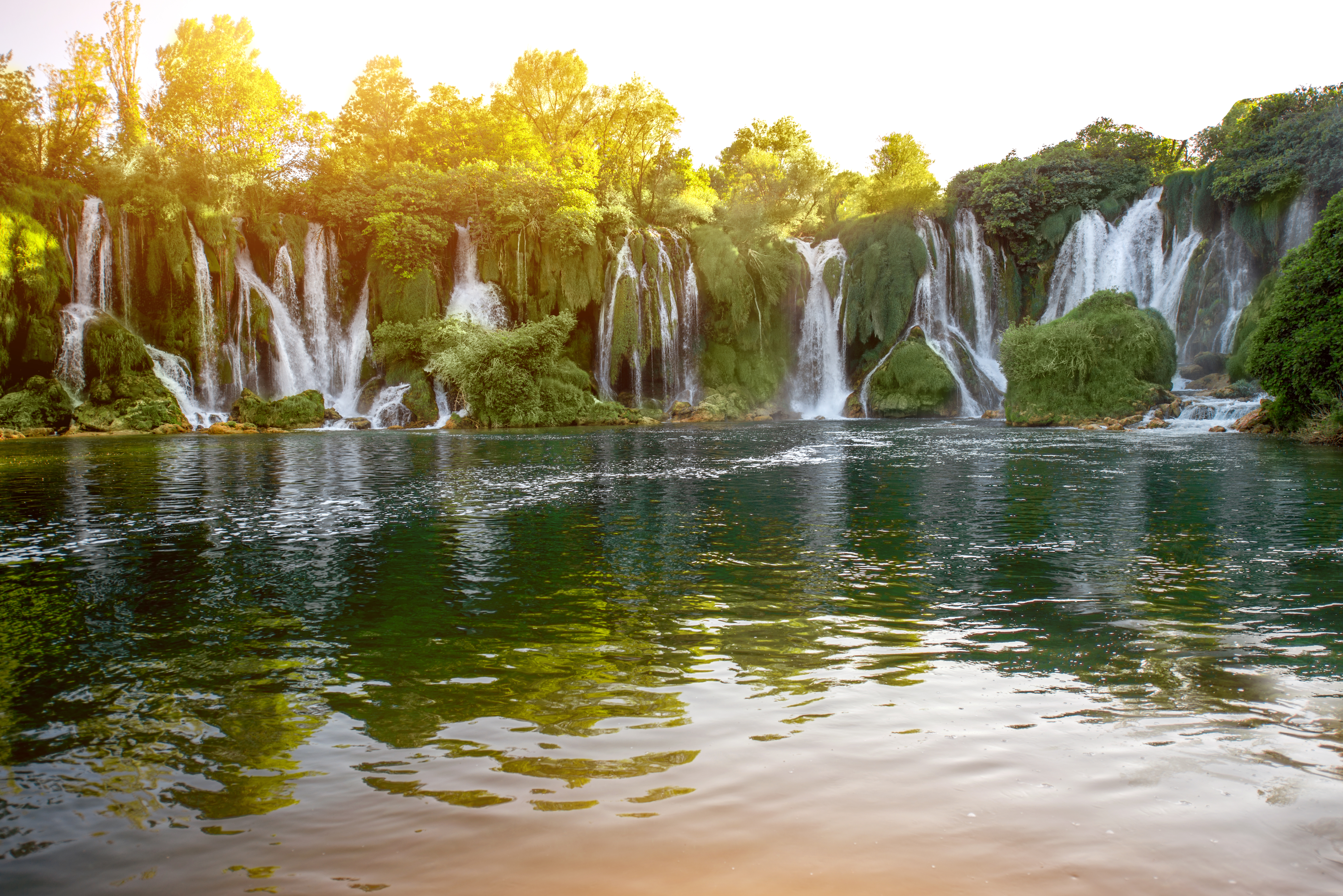 A curtain of waterfalls pours over a mossy green cliff into a wide, still pool, the sun low behind the trees.