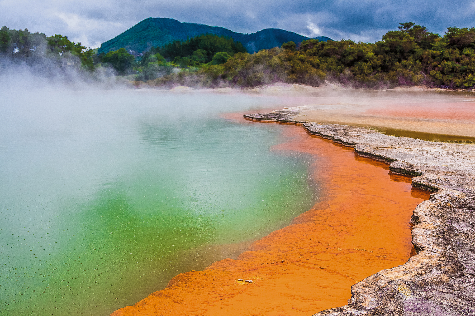 Student travelers pause beside a geothermal pool where steam rises above vivid green water and orange mineral terraces