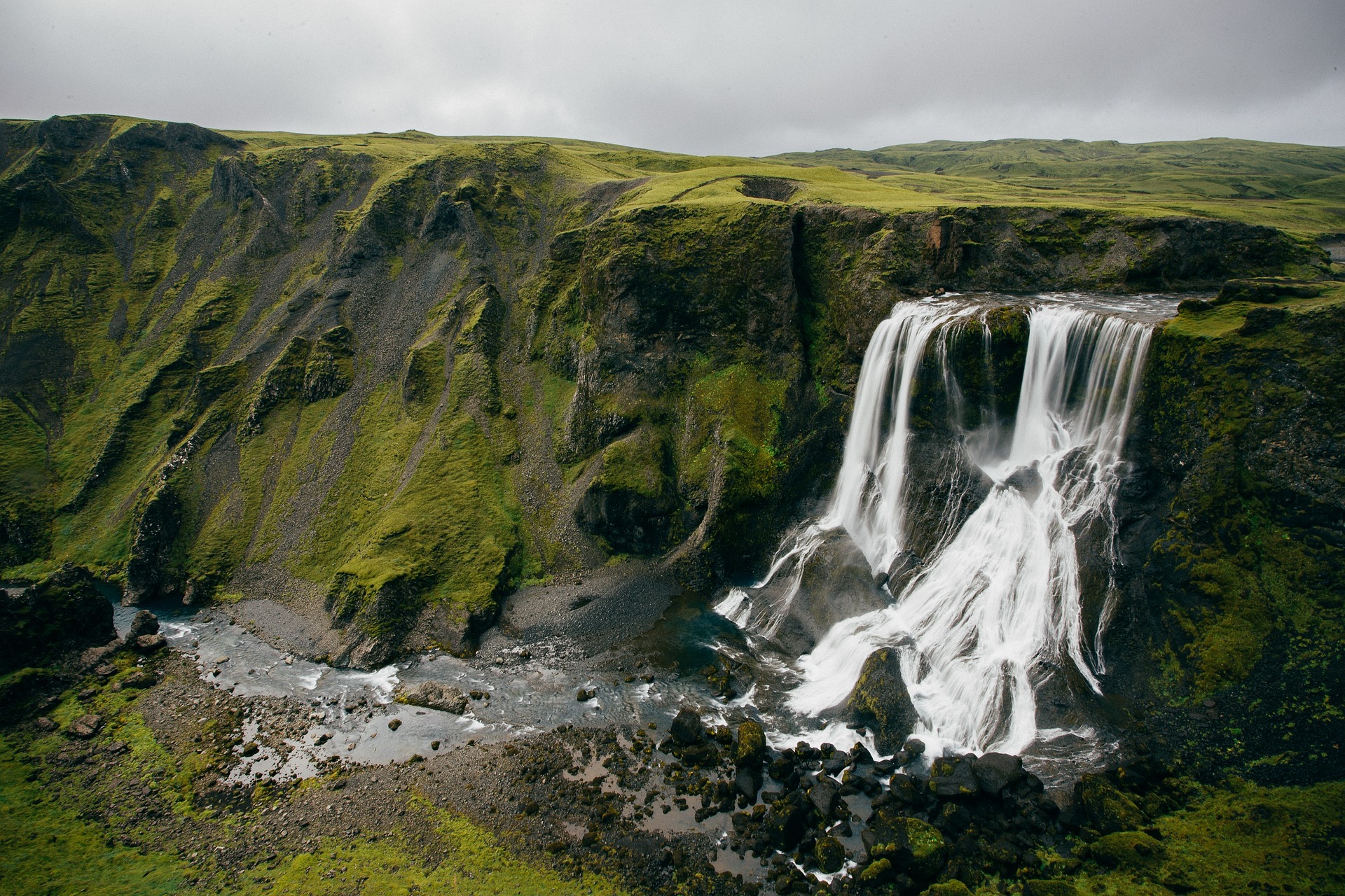 Dozens of thin waterfalls run out of the base of a mossy lava cliff and down into a river, spread along the rock face rather than falling from one point.