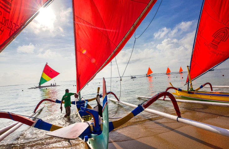 Traditional outrigger sailboats with red sails line the shore at Sanur in Bali