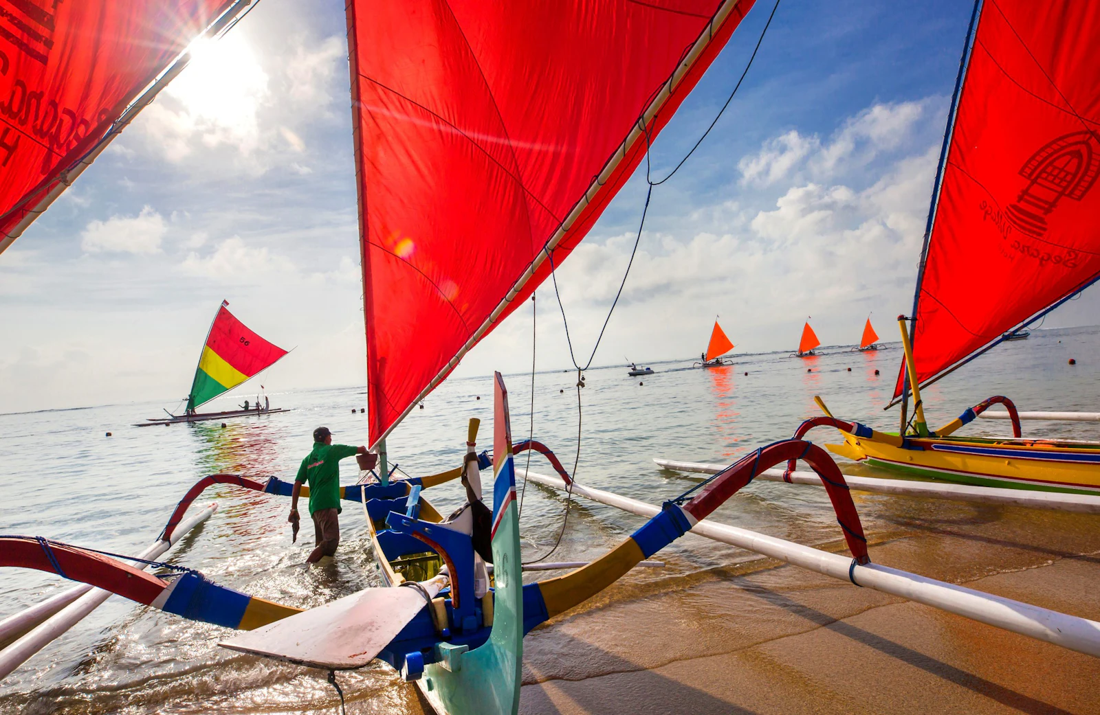 Red sails of traditional outrigger jukung boats lean over the water off Sanur beach in Bali, a boatman standing on the hull