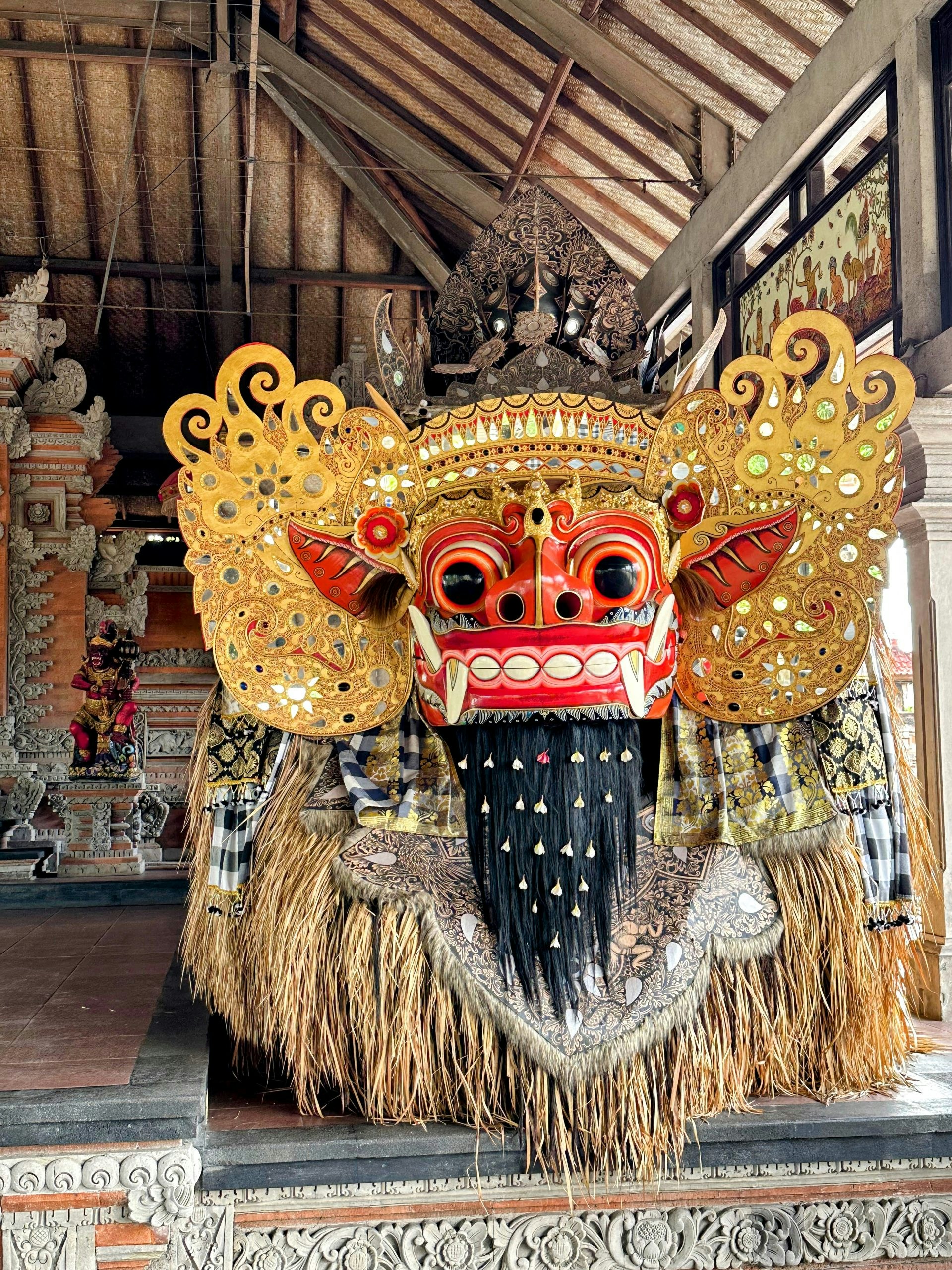 A towering ceremonial mask with a fanged red face and a gilded headdress stands inside an open wooden pavilion.
