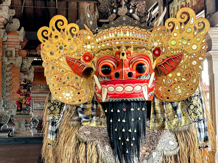 A towering ceremonial mask with a fanged red face and a gilded headdress stands inside an open wooden pavilion.