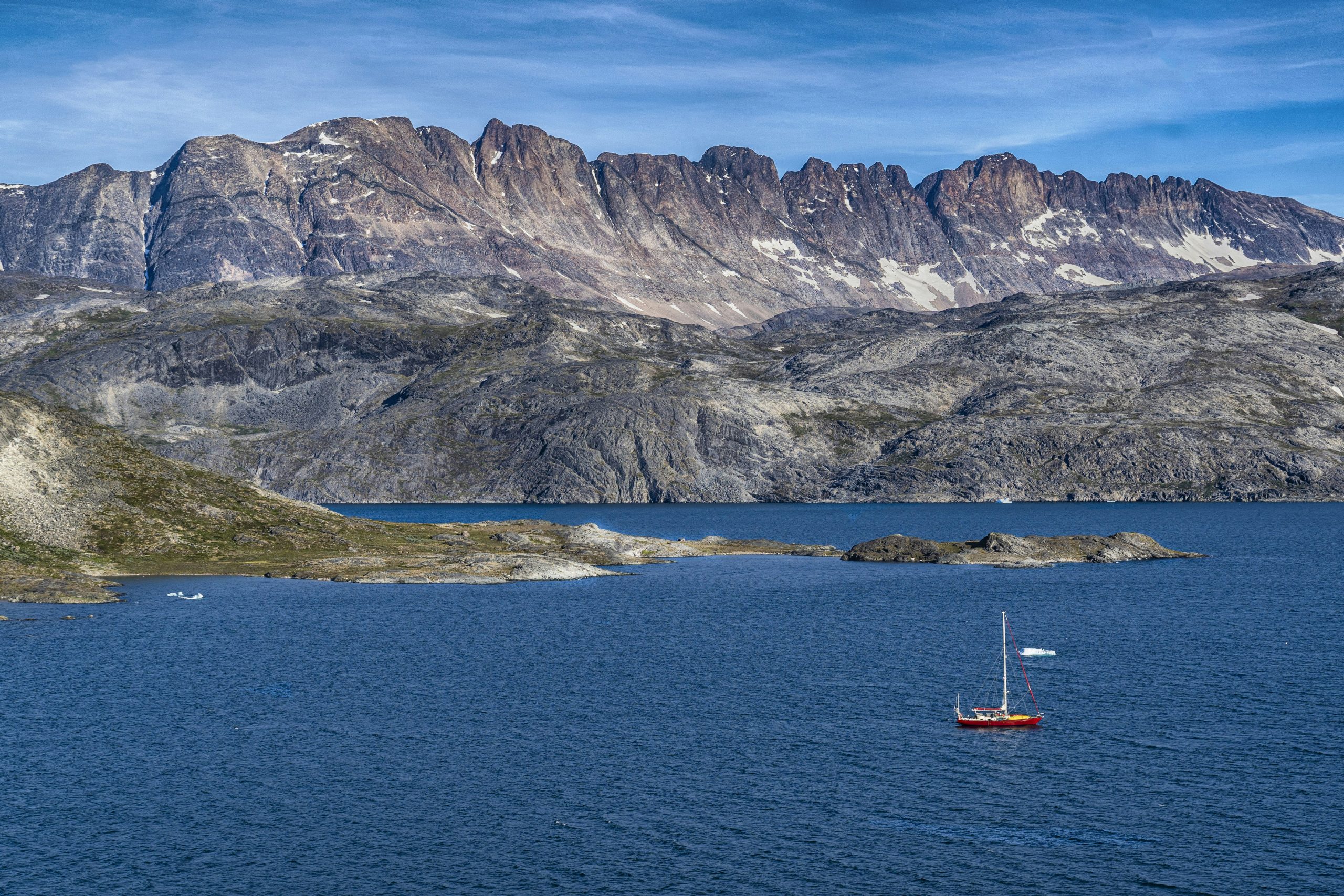 A red sailboat on a calm blue lake with a mountainous rocky terrain in the background under a clear blue sky.