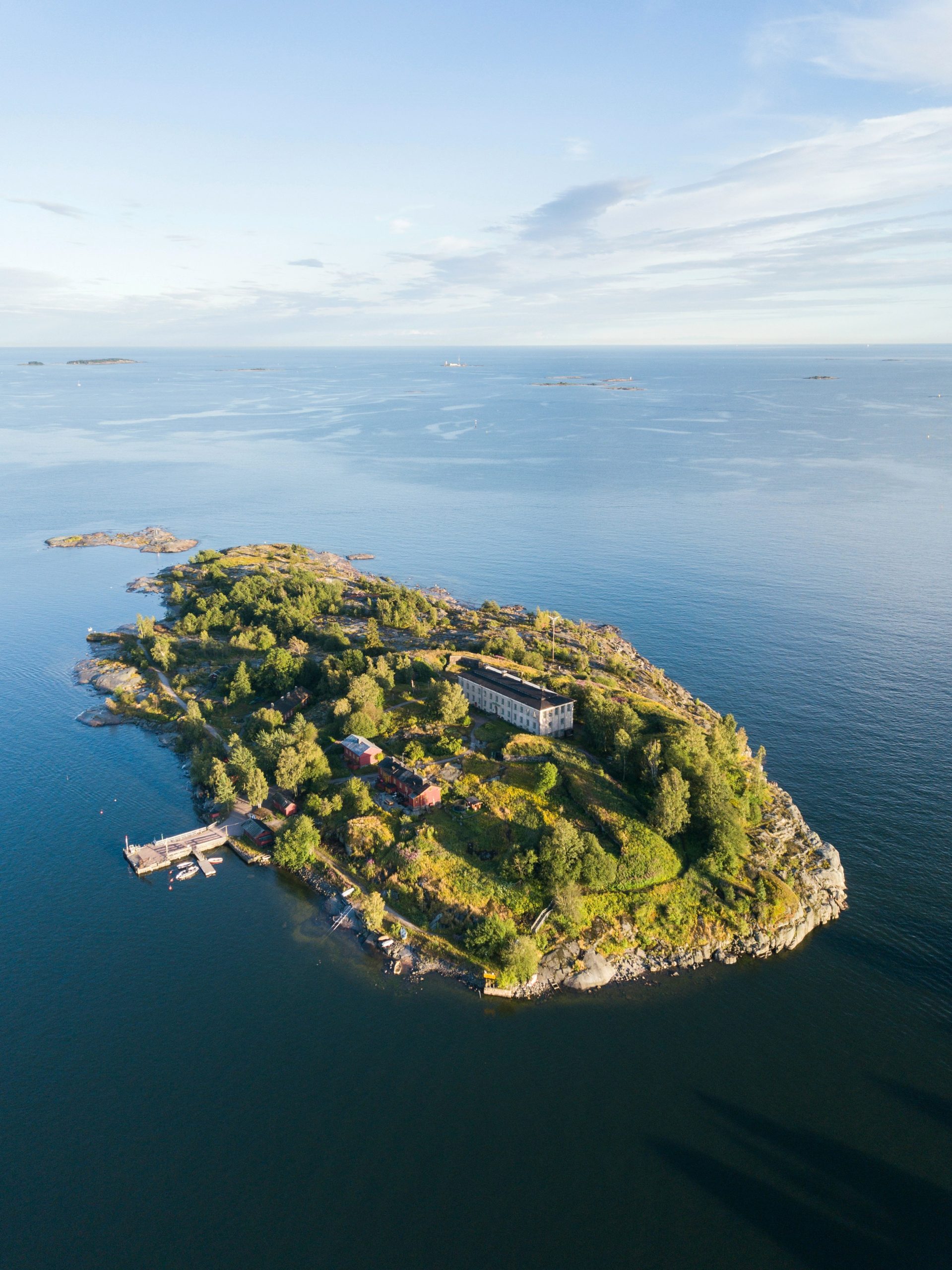 An aerial view of a small wooded island with red and white buildings and a jetty, set in calm blue water off Helsinki with more islands on the horizon.