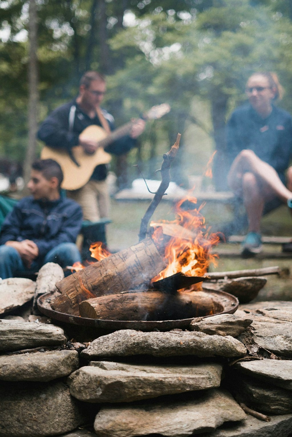 People gather around the glowing campfire, with one person strumming a guitar.