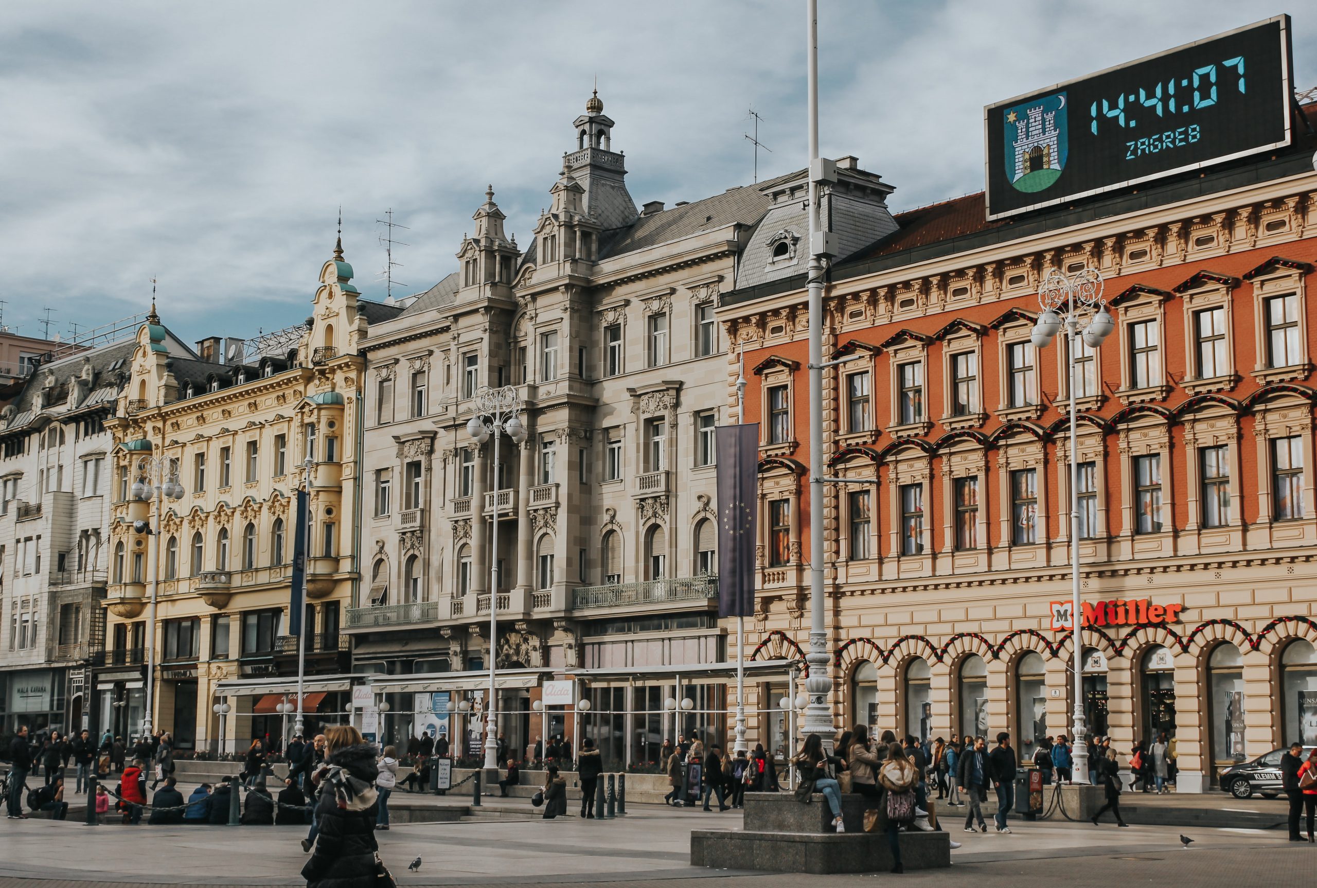 Ornate historic buildings line a plaza in Zagreb as people cross the square