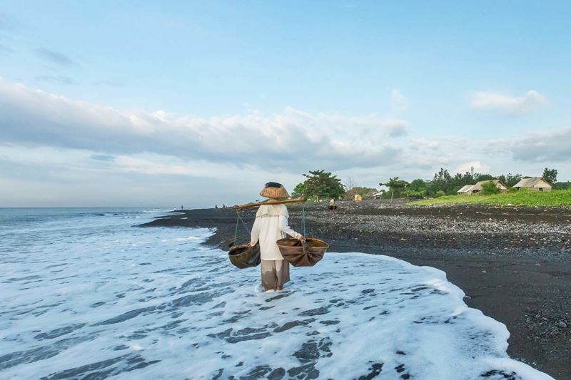 A person carries woven baskets on a pole along a black-sand beach in Indonesia with waves washing in