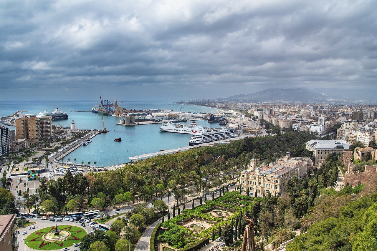 Aerial view of a coastal city harbor with cruise ships, palm-lined promenades, and gardens under a cloudy sky