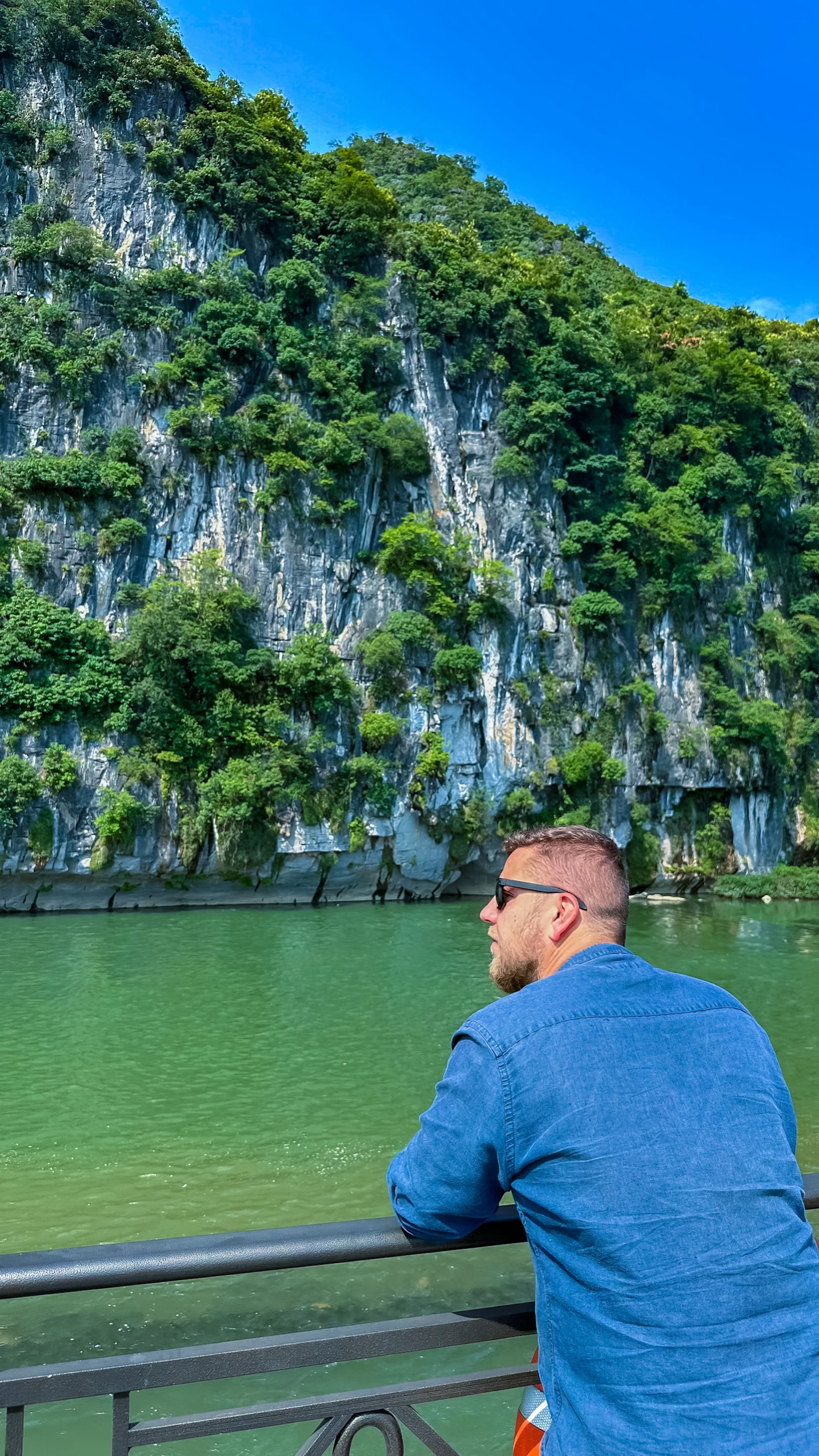 A traveler in a blue shirt looks from a boat railing toward limestone cliffs on the Li River in China