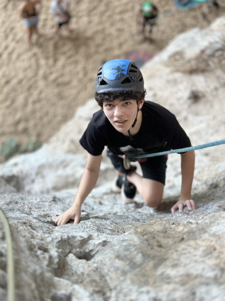 A student wearing a hardhat, attached to a rope climbing up a rockface during an outdoor program.