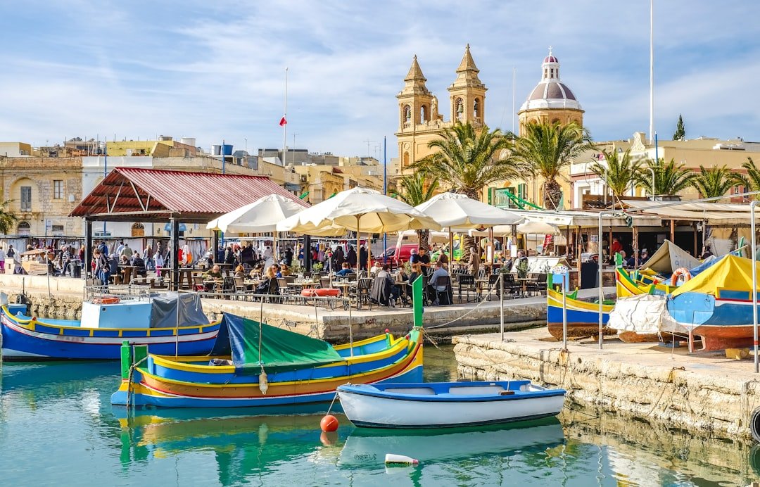Colorful traditional Maltese fishing boats moored at a harbourside market with striped awnings and a domed church behind.