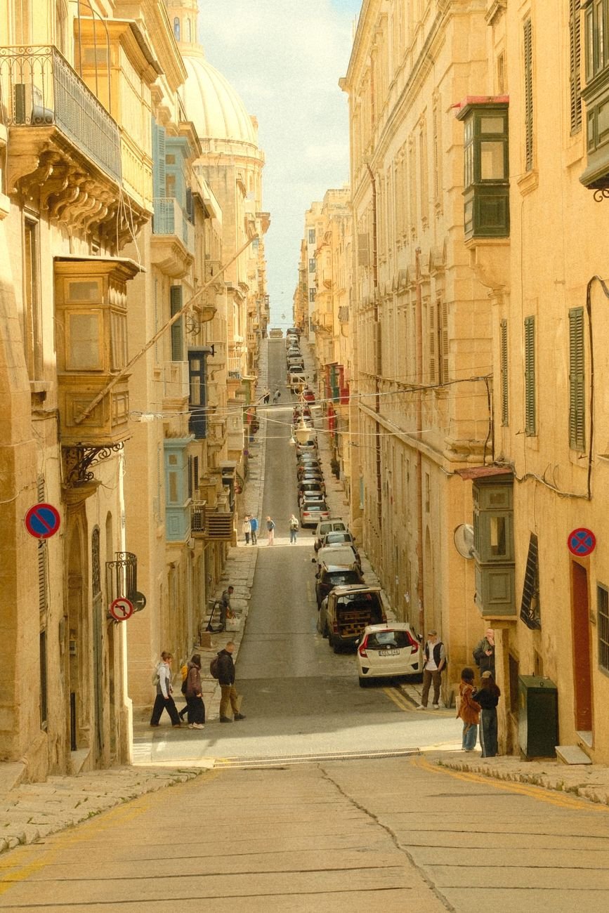 A steep, narrow street lined with sandstone buildings and wooden balconies, with a cathedral dome in the distance.