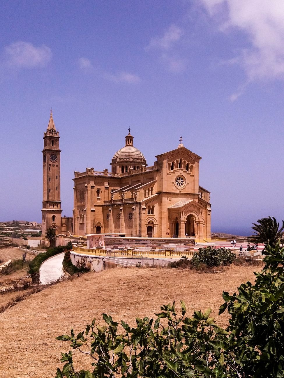 A honey-colored hilltop basilica with a tall bell tower, seen from its plaza among farm fields.