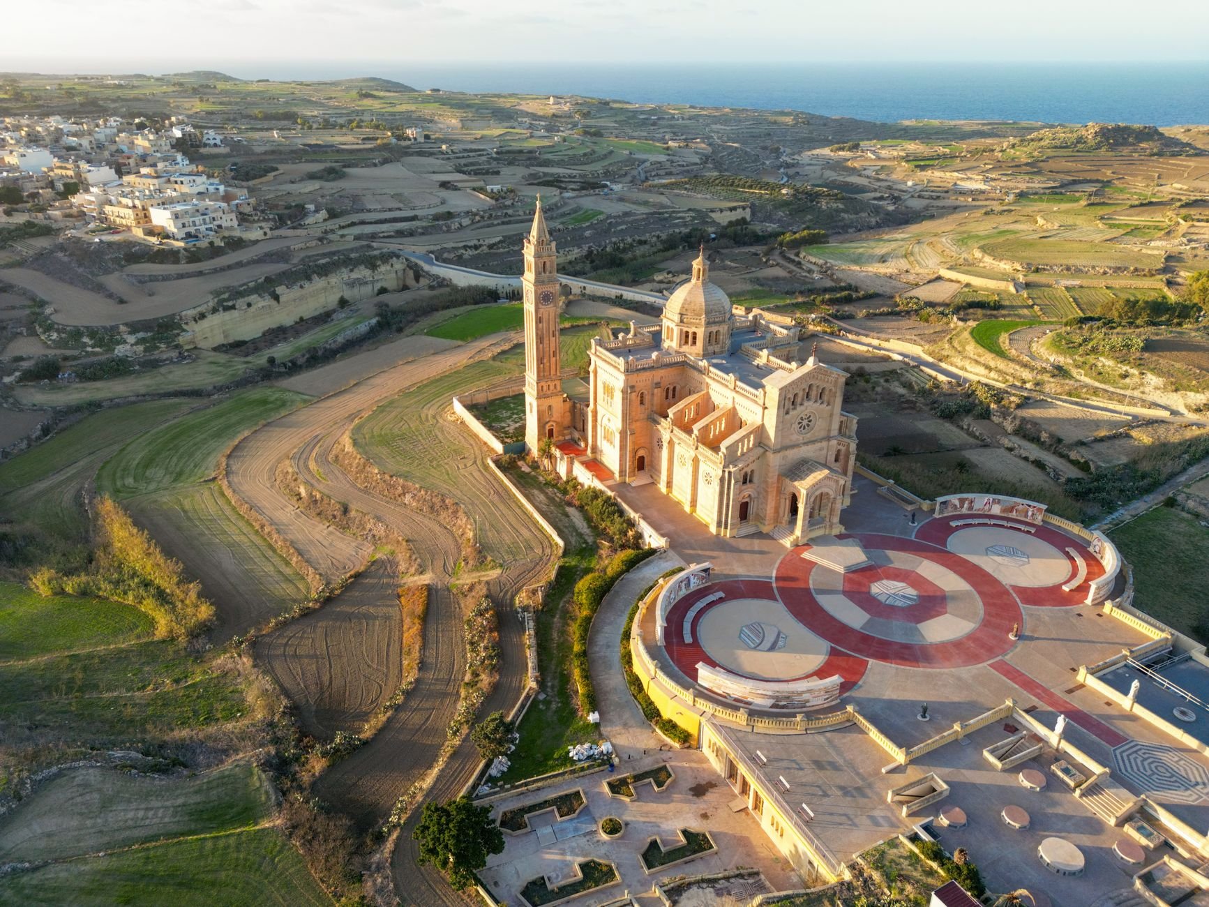 An aerial view of a hilltop basilica with a tall bell tower and dome, rising above patchwork farmland near the coast.