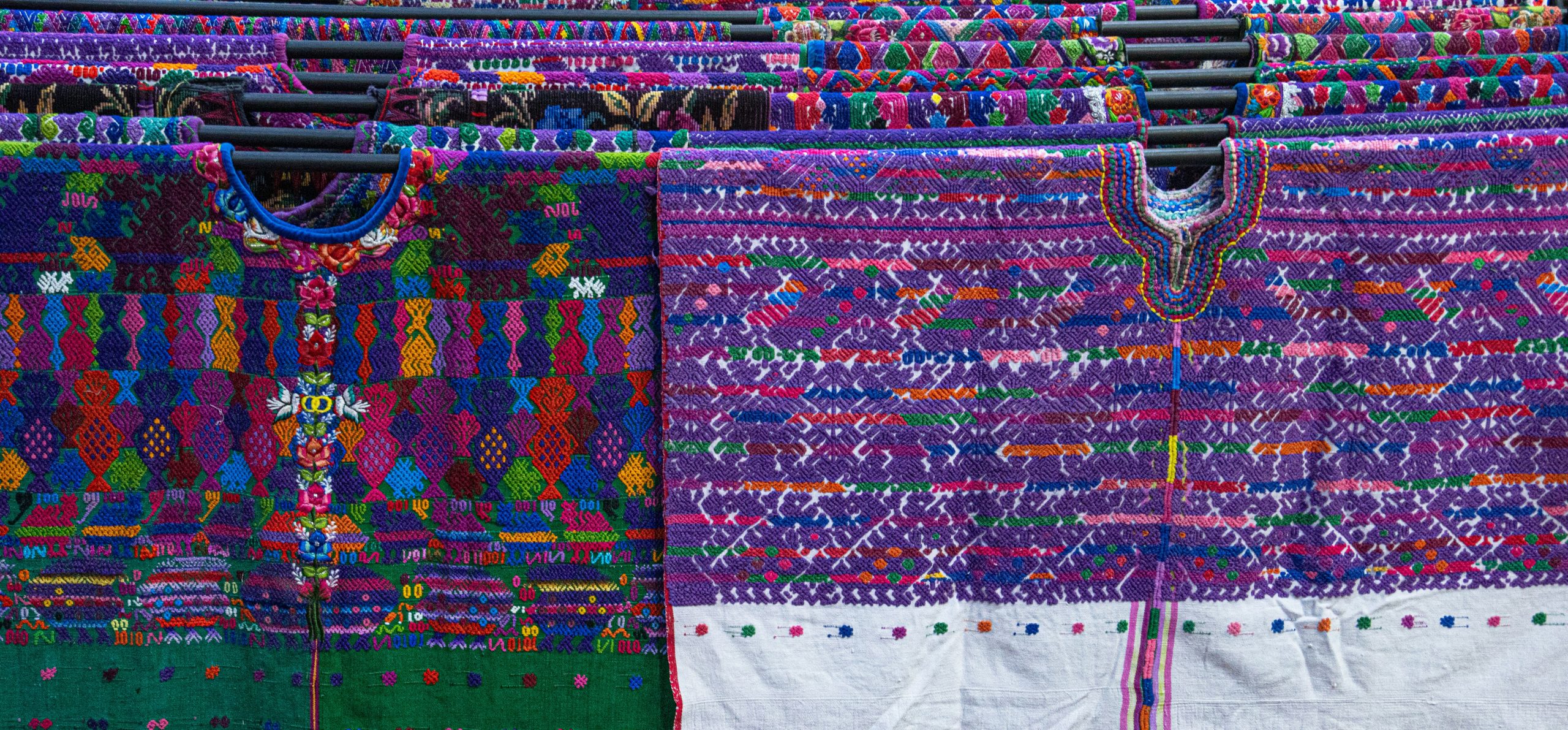 Hand-woven blouses hung on a rail, dense with brocade figures in purple, green and red over white cloth.
