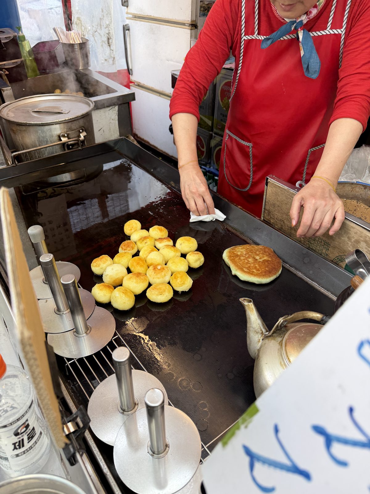 A vendor in a red apron wipes a griddle of frying hotteok at a stall in Mangwon Market, Seoul