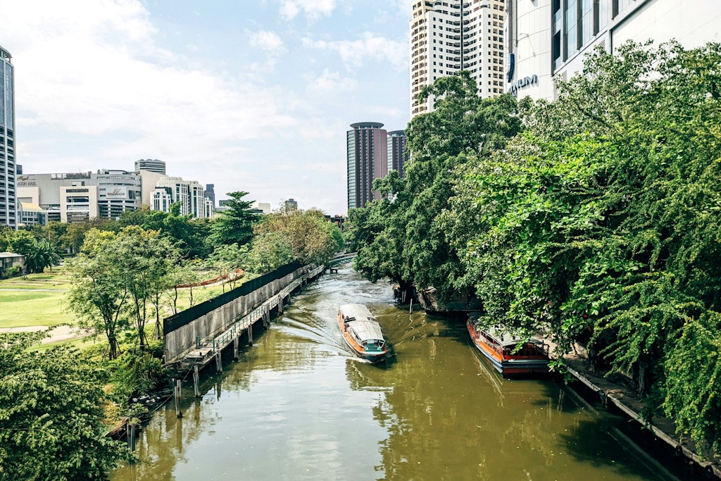 A calm canal in Bangkok is surrounded by green plants and trees. A small boat floats on the water. The area feels peaceful, offering a break from the busy city life.