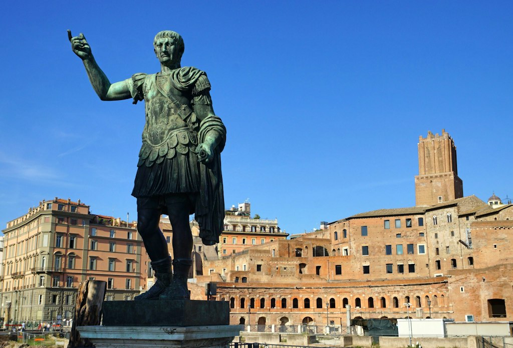 Bronze statue of Emperor Trajan standing tall, set against the backdrop of ancient Roman ruins under a clear blue sky.