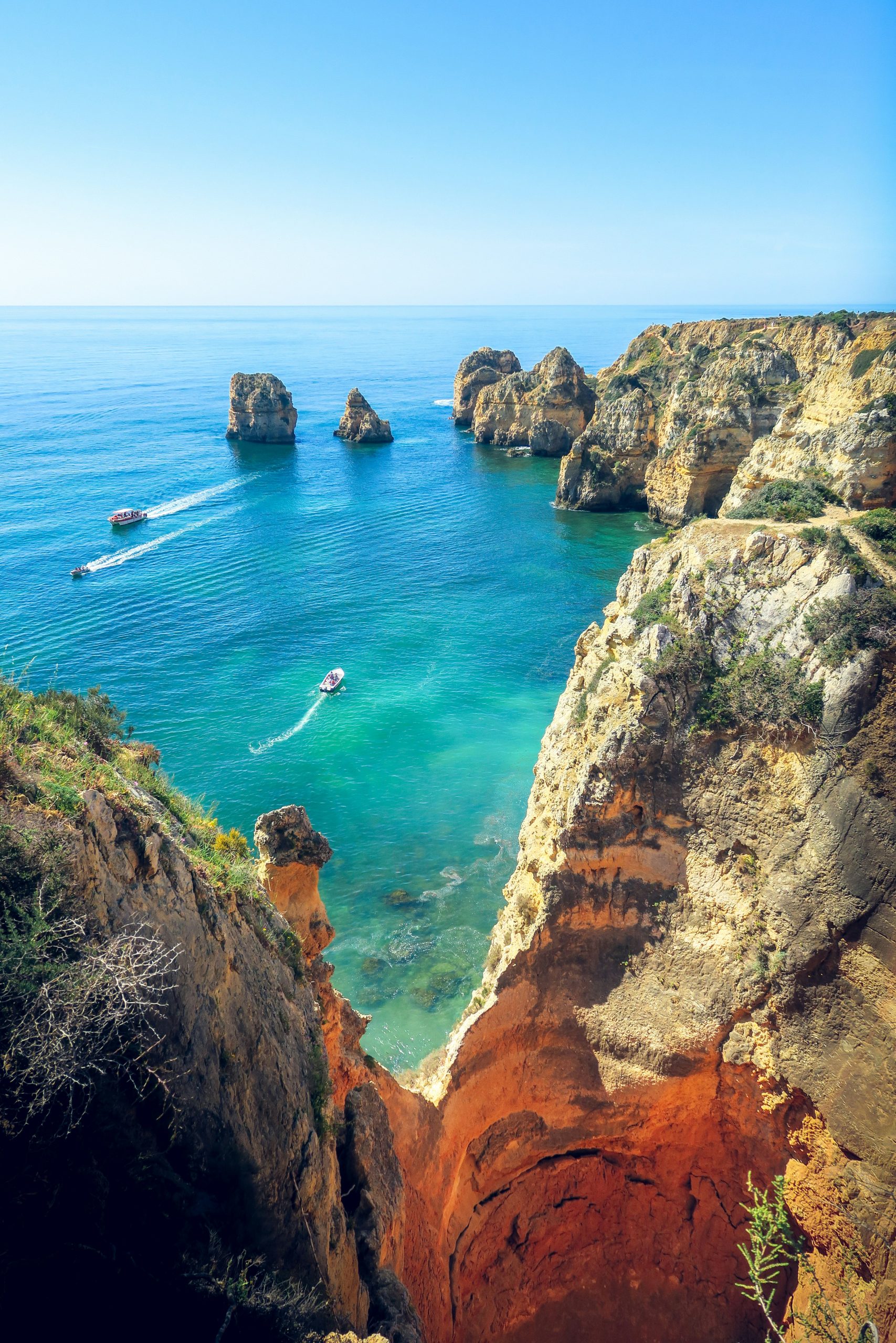 A breathtaking view of a rocky coastline with steep cliffs overlooking clear turquoise waters. A small boat speeds across the sea, leaving a white trail behind. fun facts about portugal