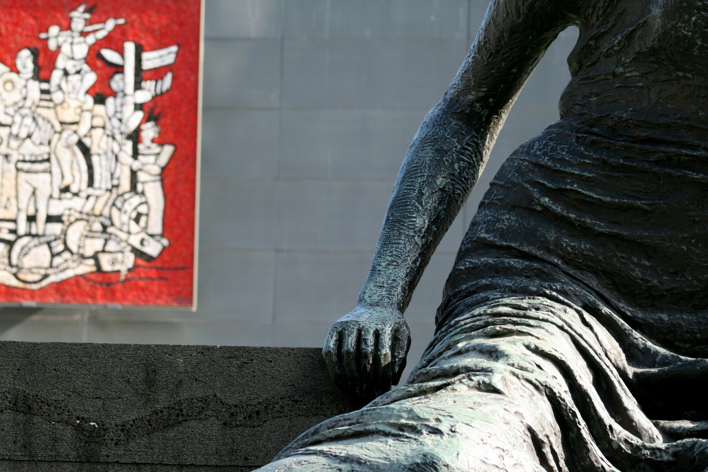 A weathered bronze figure seated outside a gallery wall hung with a red artwork