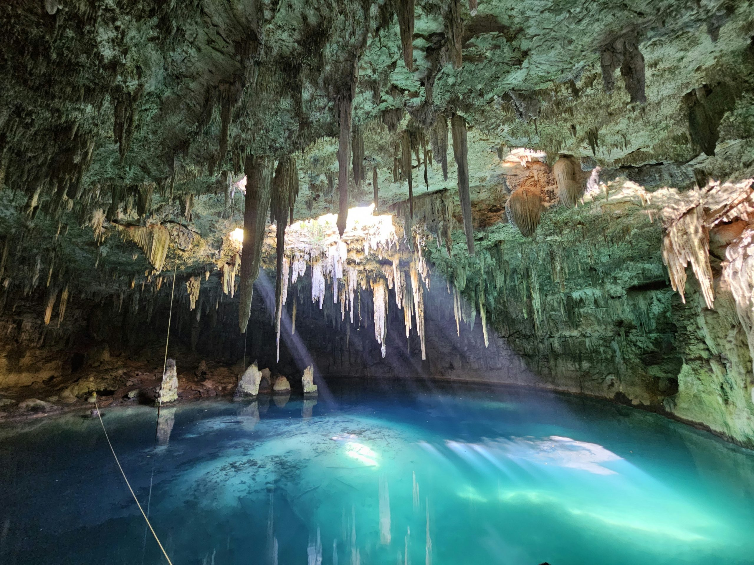 A closed cenote: a cavern of stalactites over still turquoise water, with daylight falling through an opening in the roof.