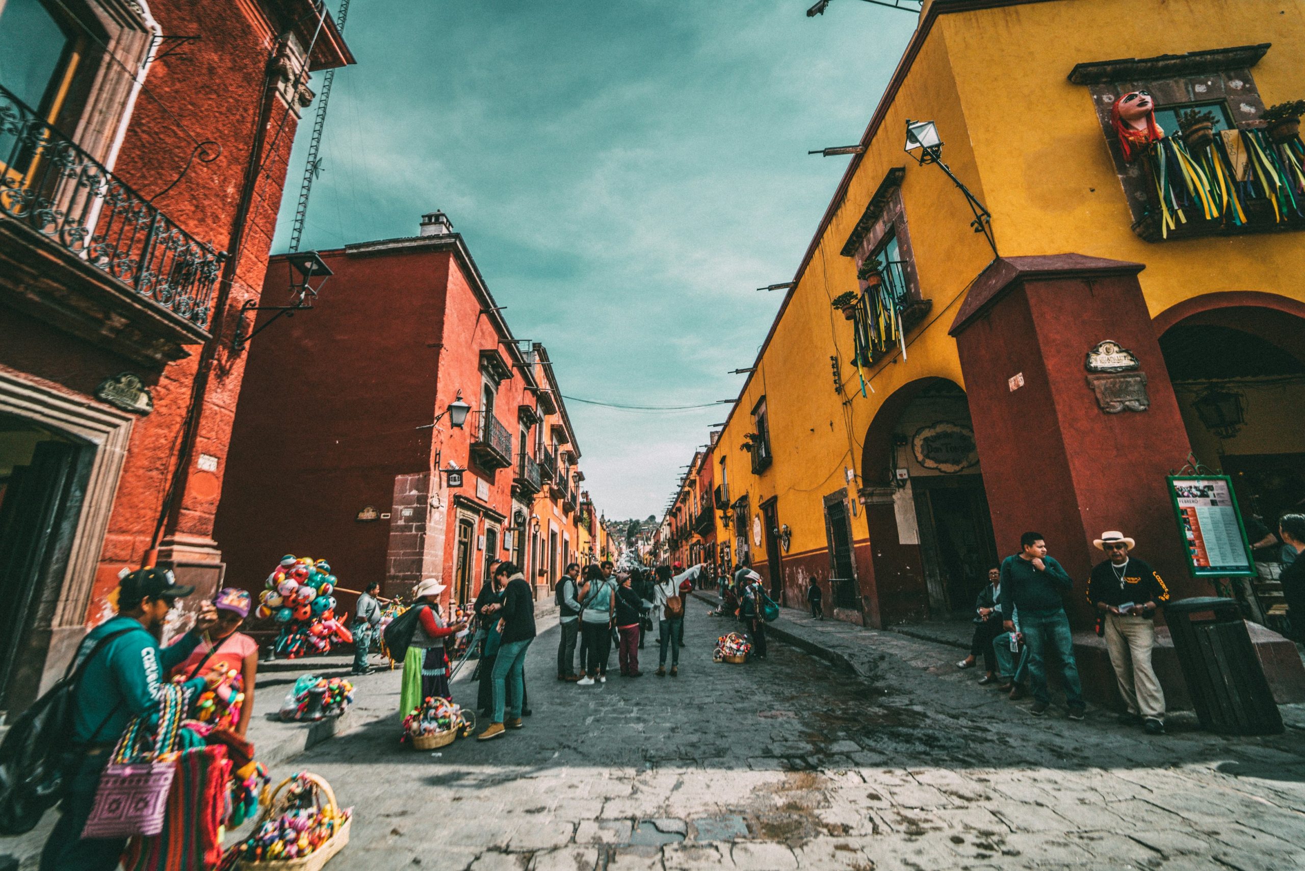 A steep cobbled street between ochre and terracotta colonial buildings, with vendors selling balloons and woven goods and hills visible at the far end.