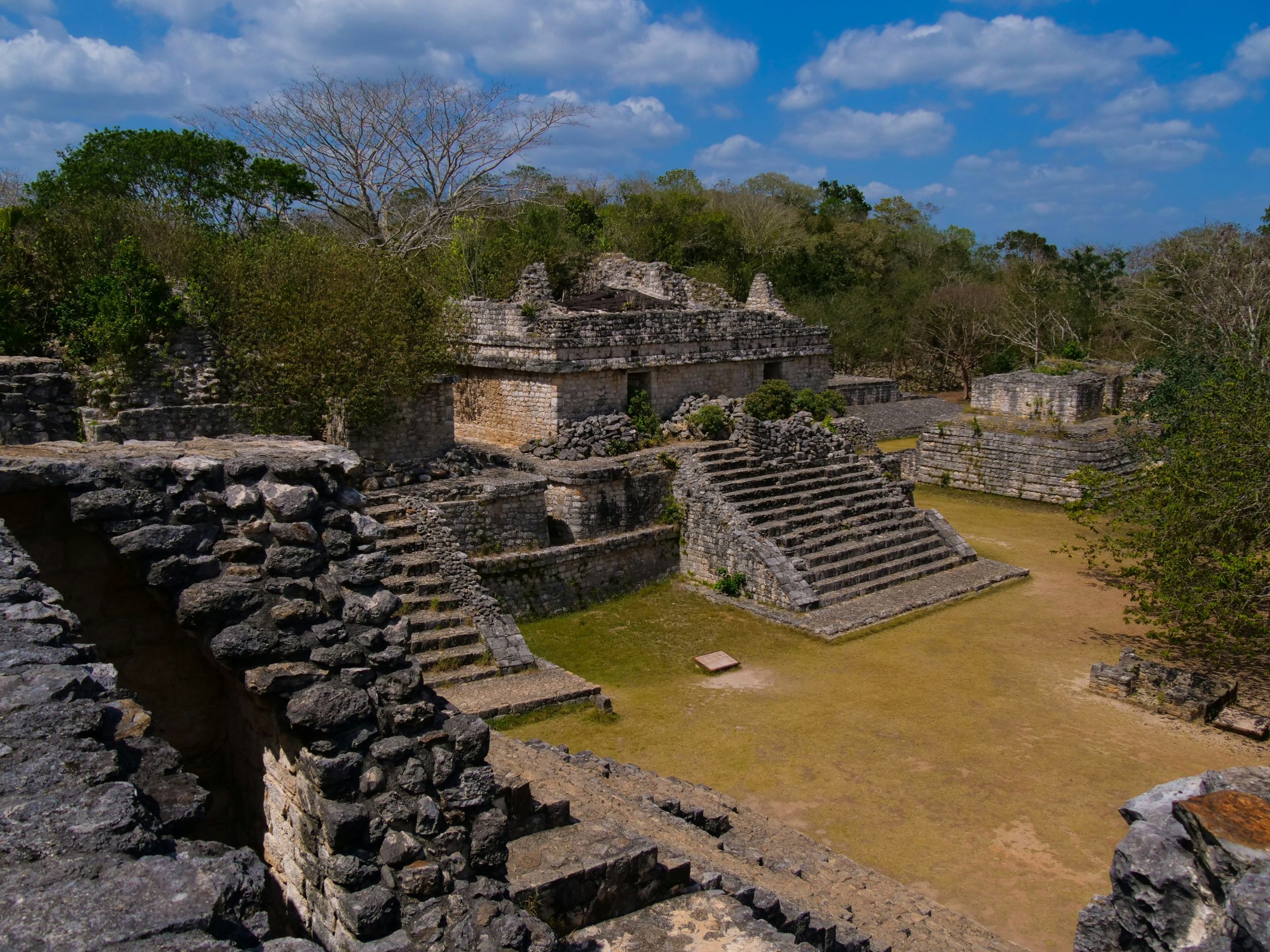 A walled Maya plaza of limestone buildings and stepped platforms, surrounded by low dry jungle.