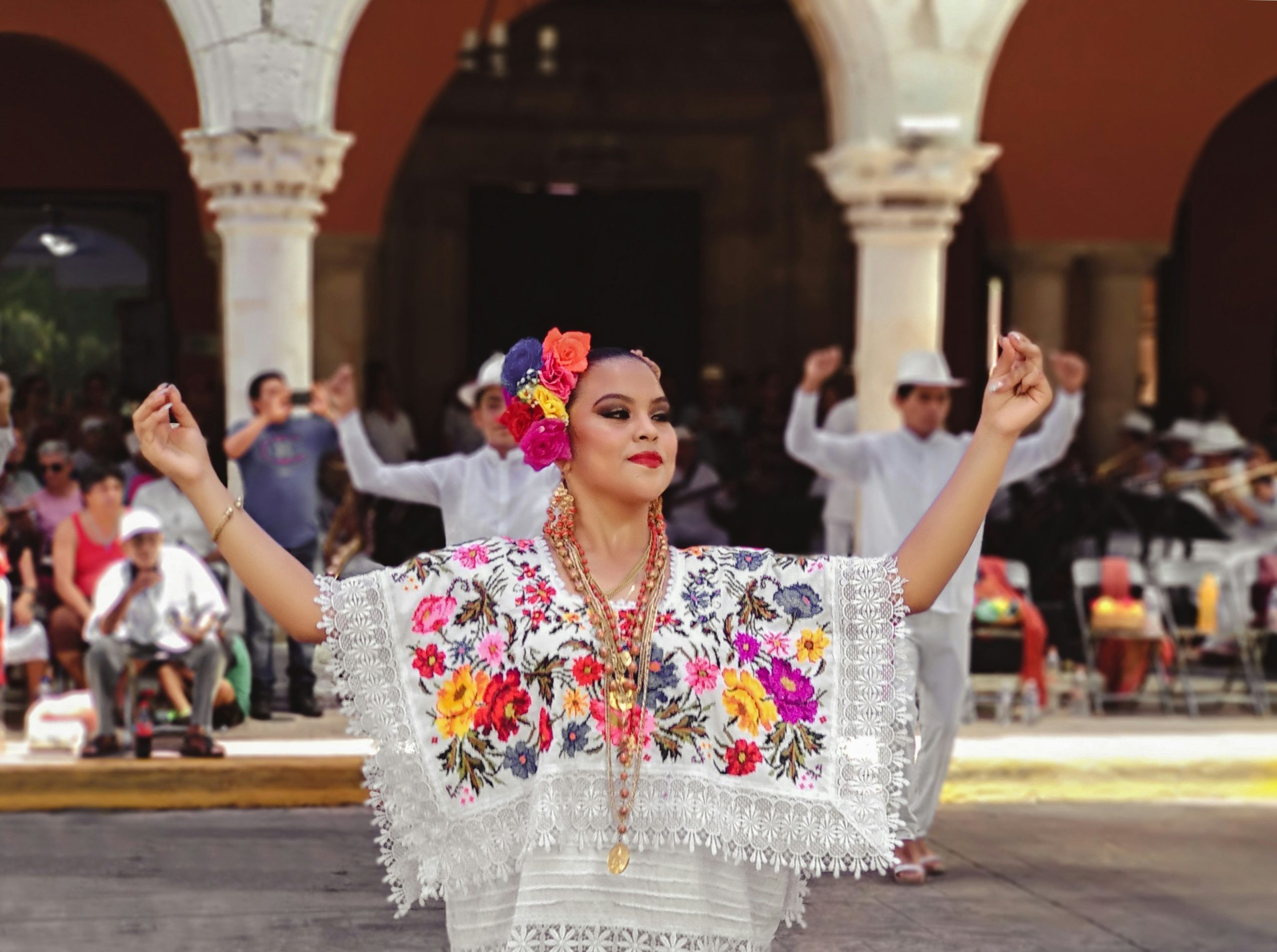 A dancer in a white embroidered dress with flowers in her hair, arms raised mid-step, with musicians and other dancers under stone arches behind her.
