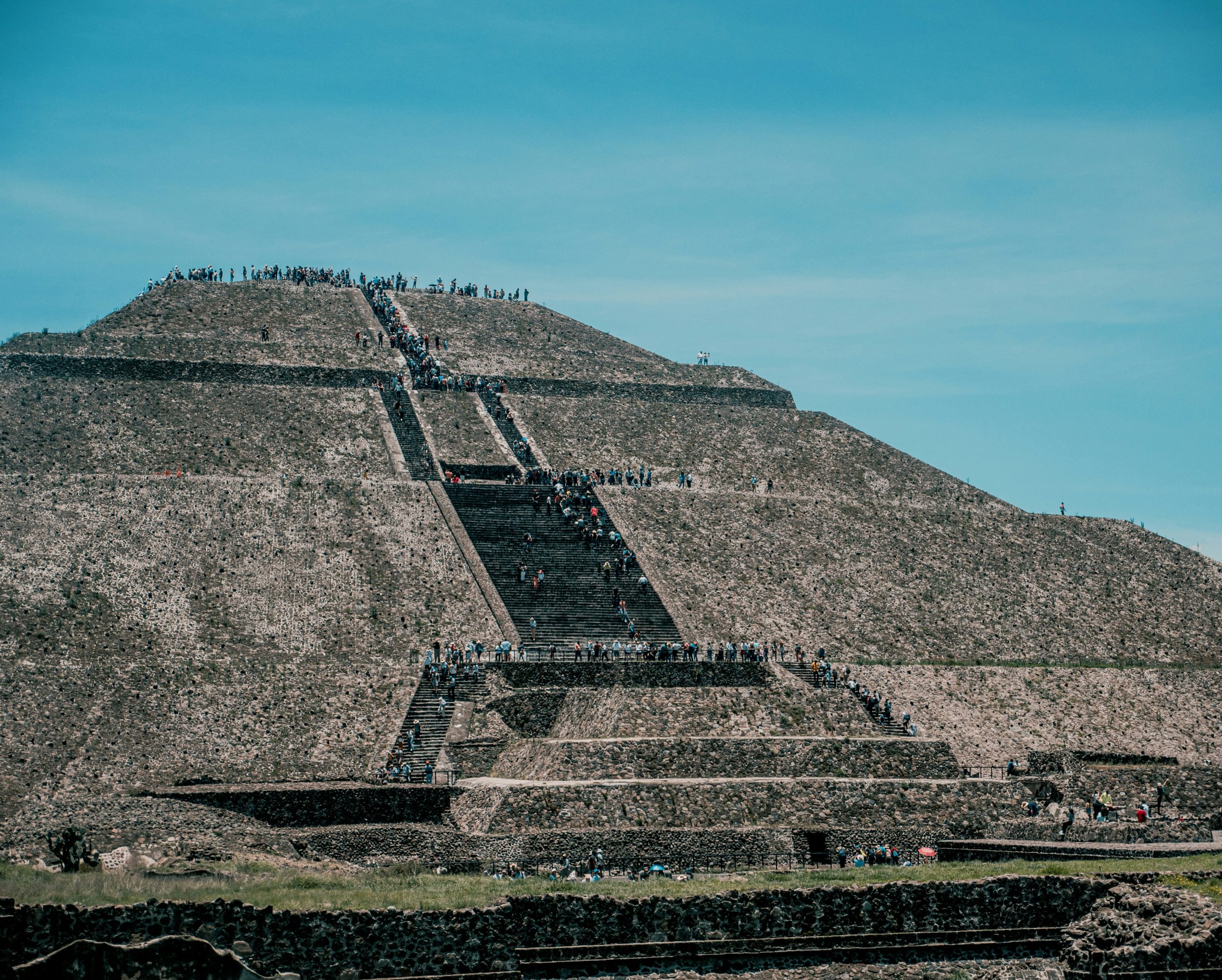 The stepped face and central stairway of a large stone pyramid, with visitors climbing it in a line.