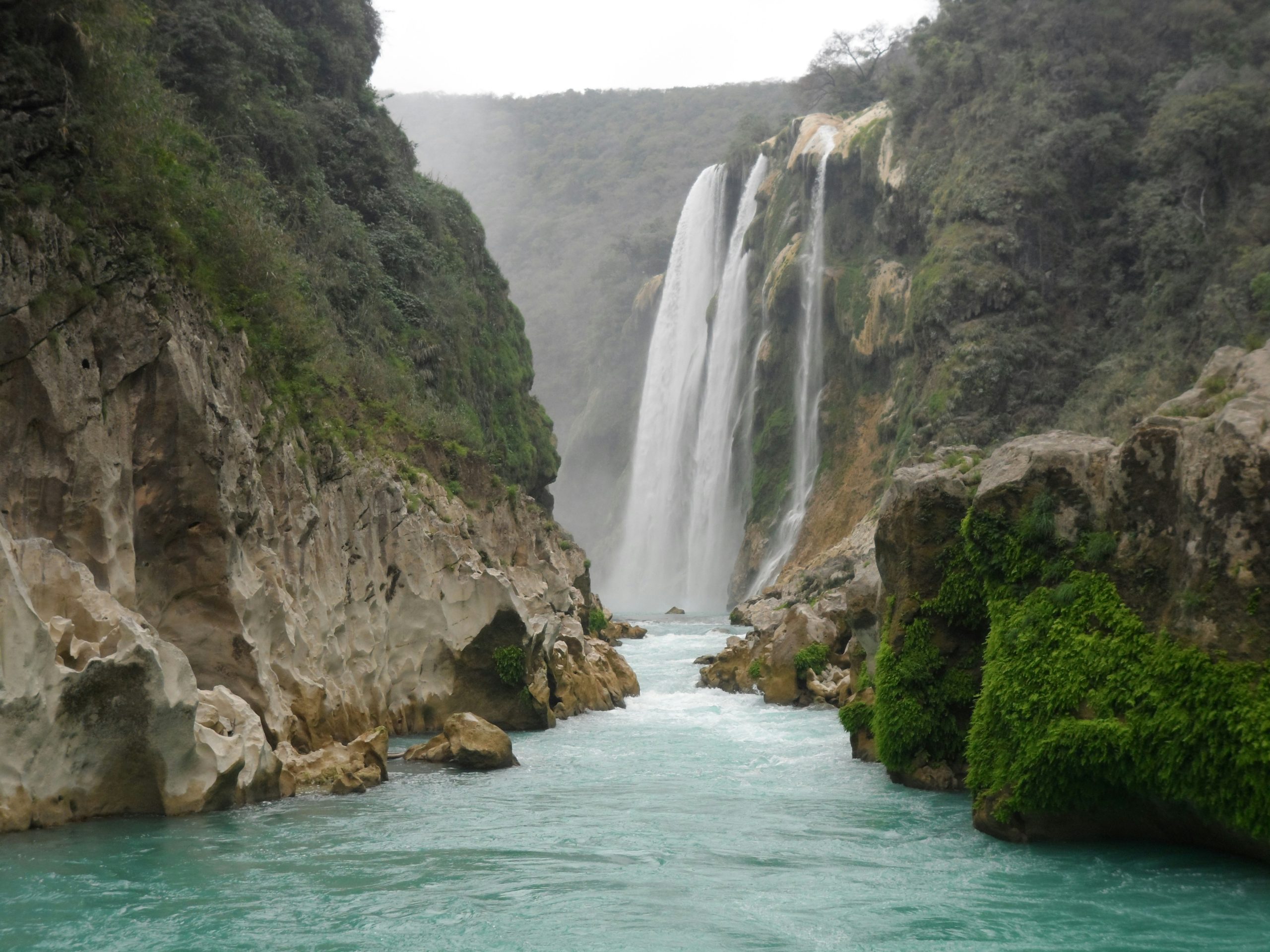 A wide waterfall dropping into a turquoise river gorge between limestone cliffs hung with greenery.