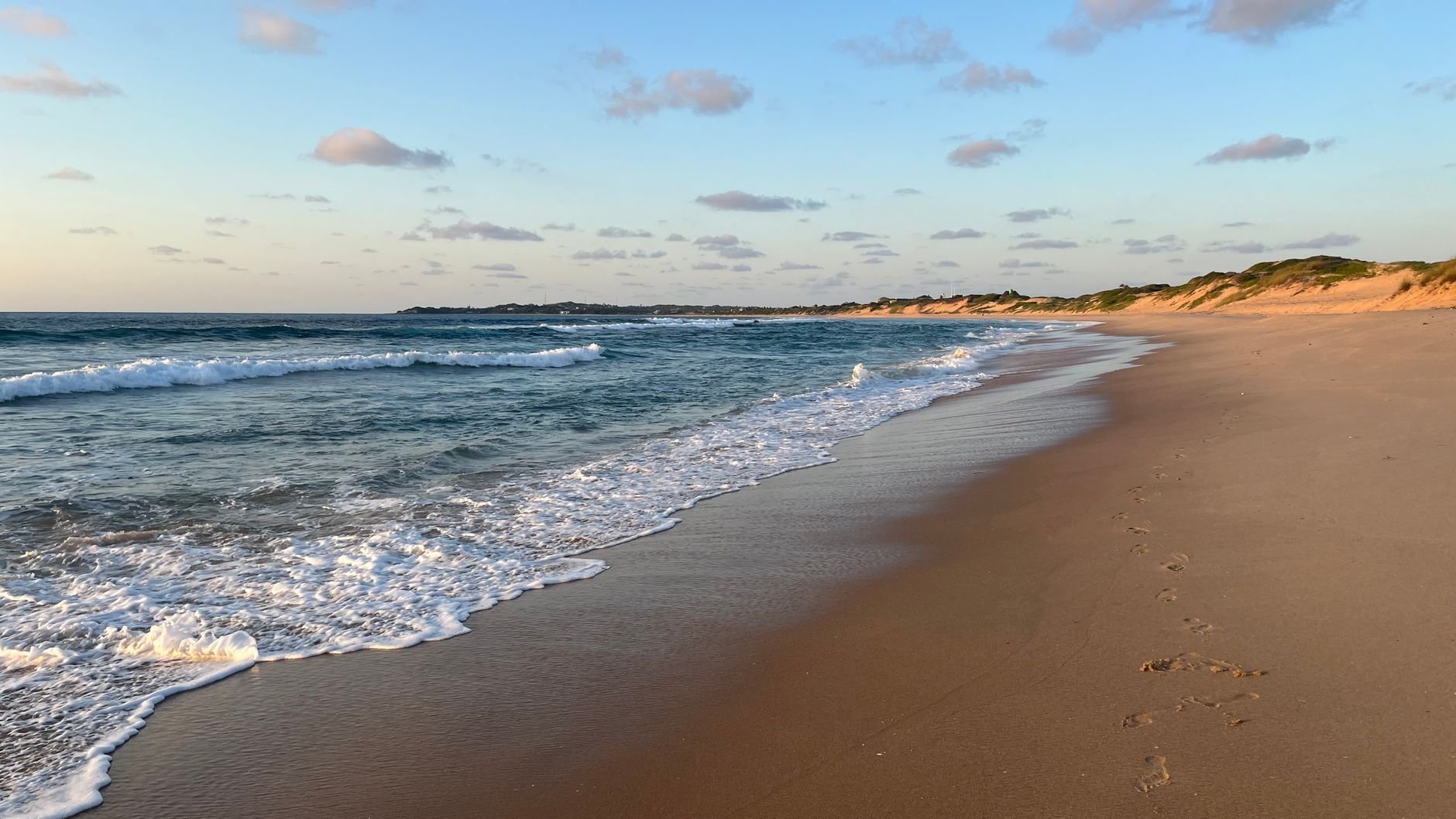 Footprints lead along a wide sandy beach at sunset, with gentle waves, and a headland in the distance.
